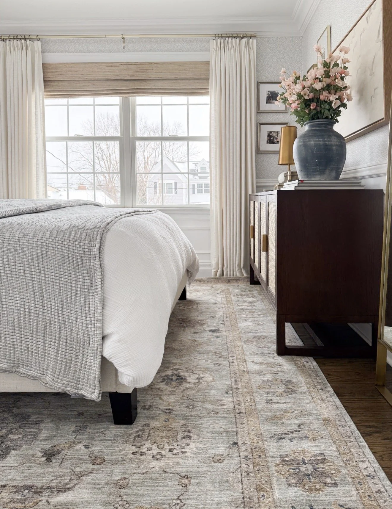 Bright and airy bedroom featuring a bed with gray bedding and a light gray blanket, styled with a dark wood dresser with cane detailing, topped with a blue vase of pink flowers and decorative books, next to tall windows with cream curtains.