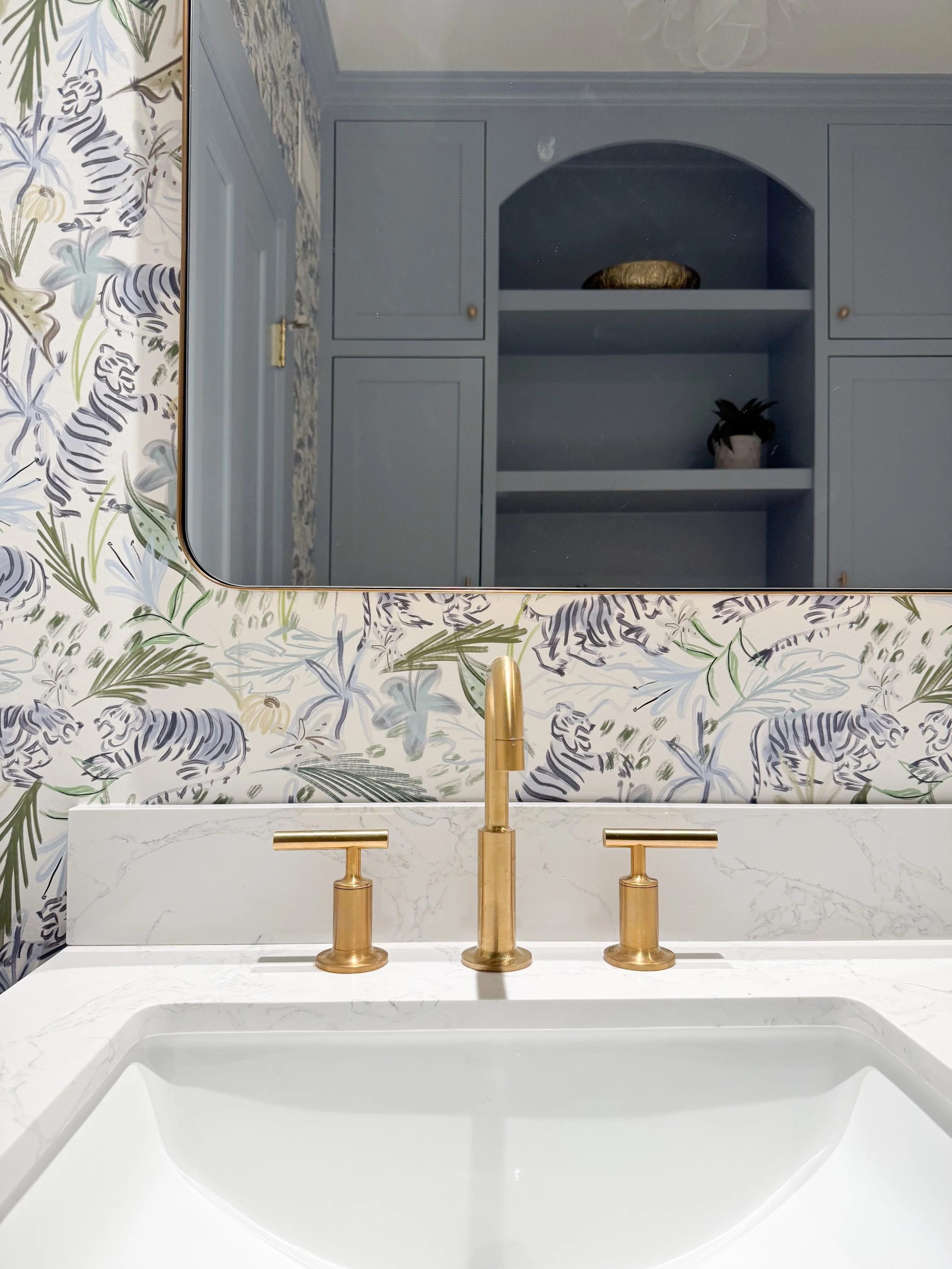 Brass bathroom faucet and round brass mirror paired with blue tiger wallpaper and a marble vanity countertop.