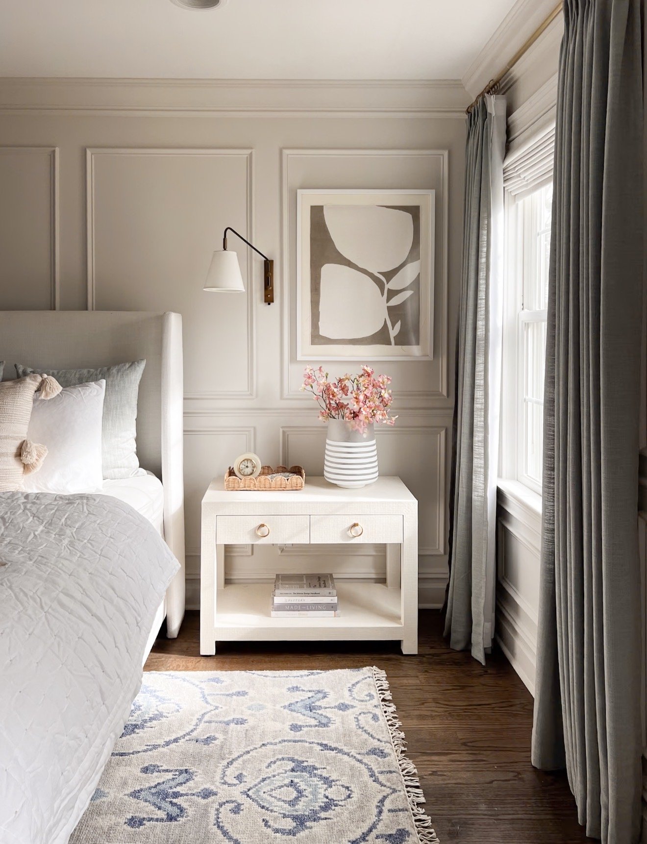 Serena & Lily Driftway bedside table in a neutral primary bedroom with white bedding and a striped gray throw on the bed, natural light from windows highlighting the tabletop decor with books and a vase of flowers