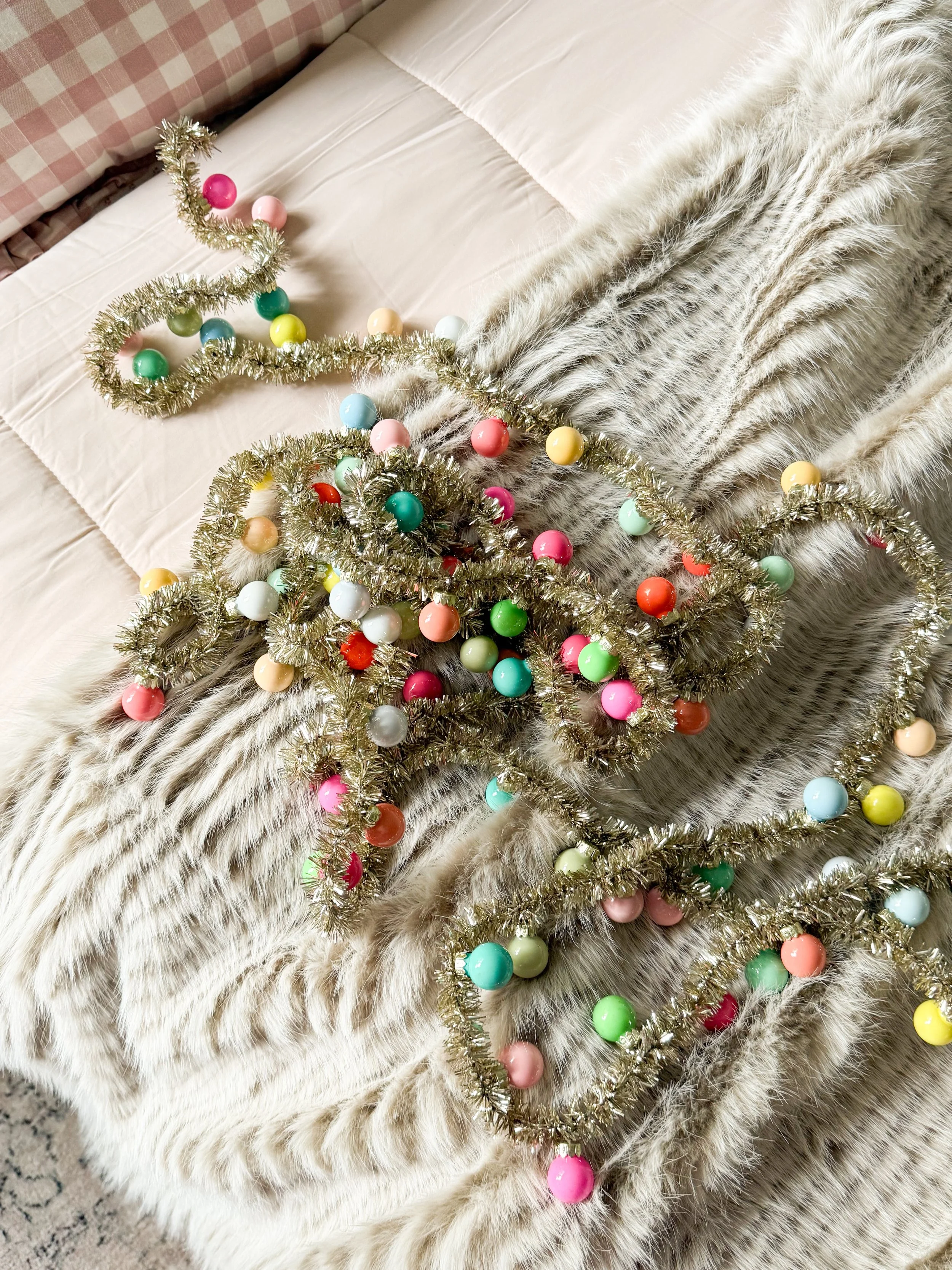 Anthropologie holiday decor featuring colorful vintage inspired Christmas garland with gold tinsel and multicolored baubles draped across a plush faux fur throw on a bed with blush pink bedding.