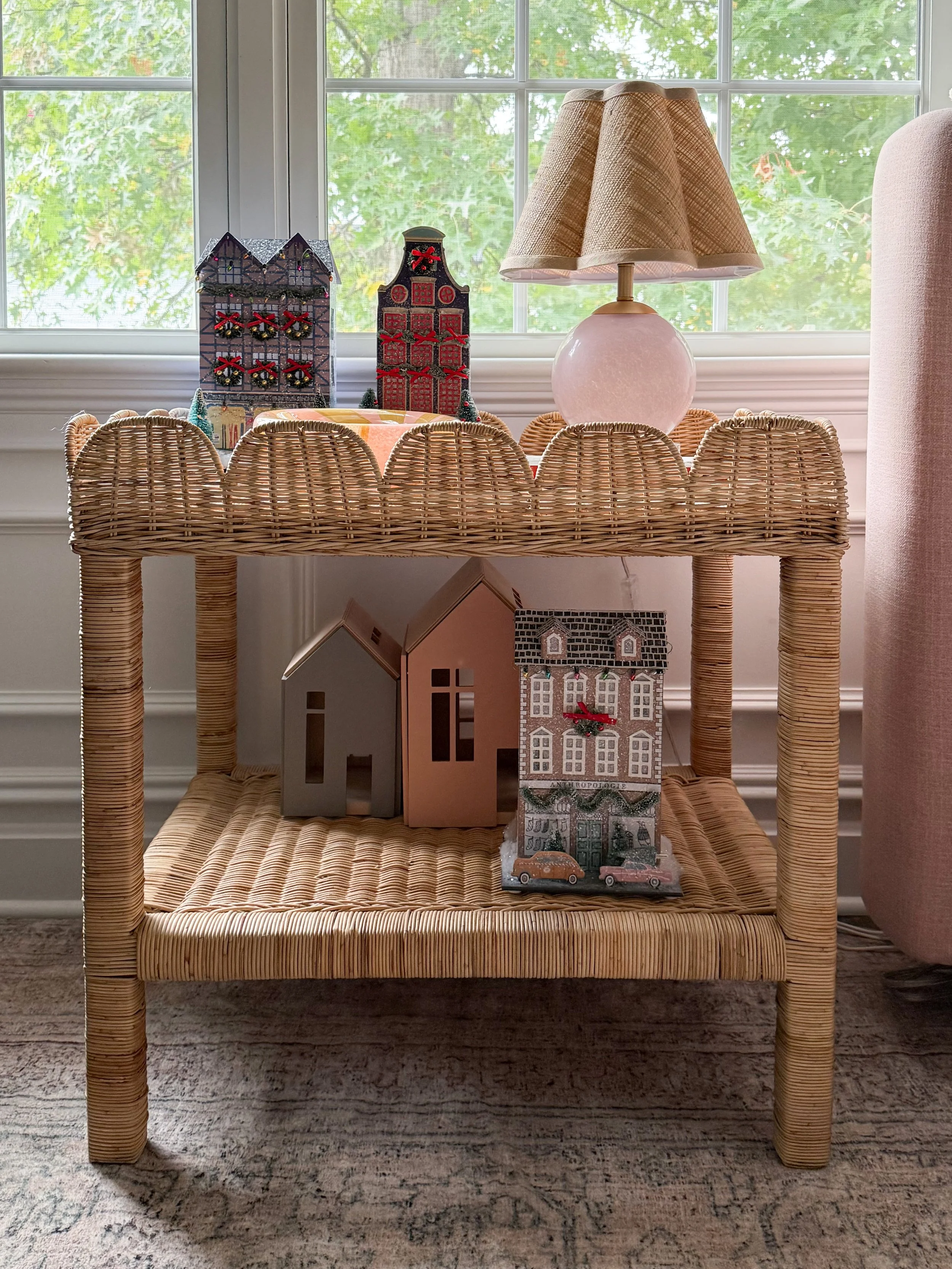 Anthropologie holiday decor featuring miniature Christmas village houses with red bows, a pink glass table lamp with a woven shade, and a colorful decorative dish displayed on a rattan side table.