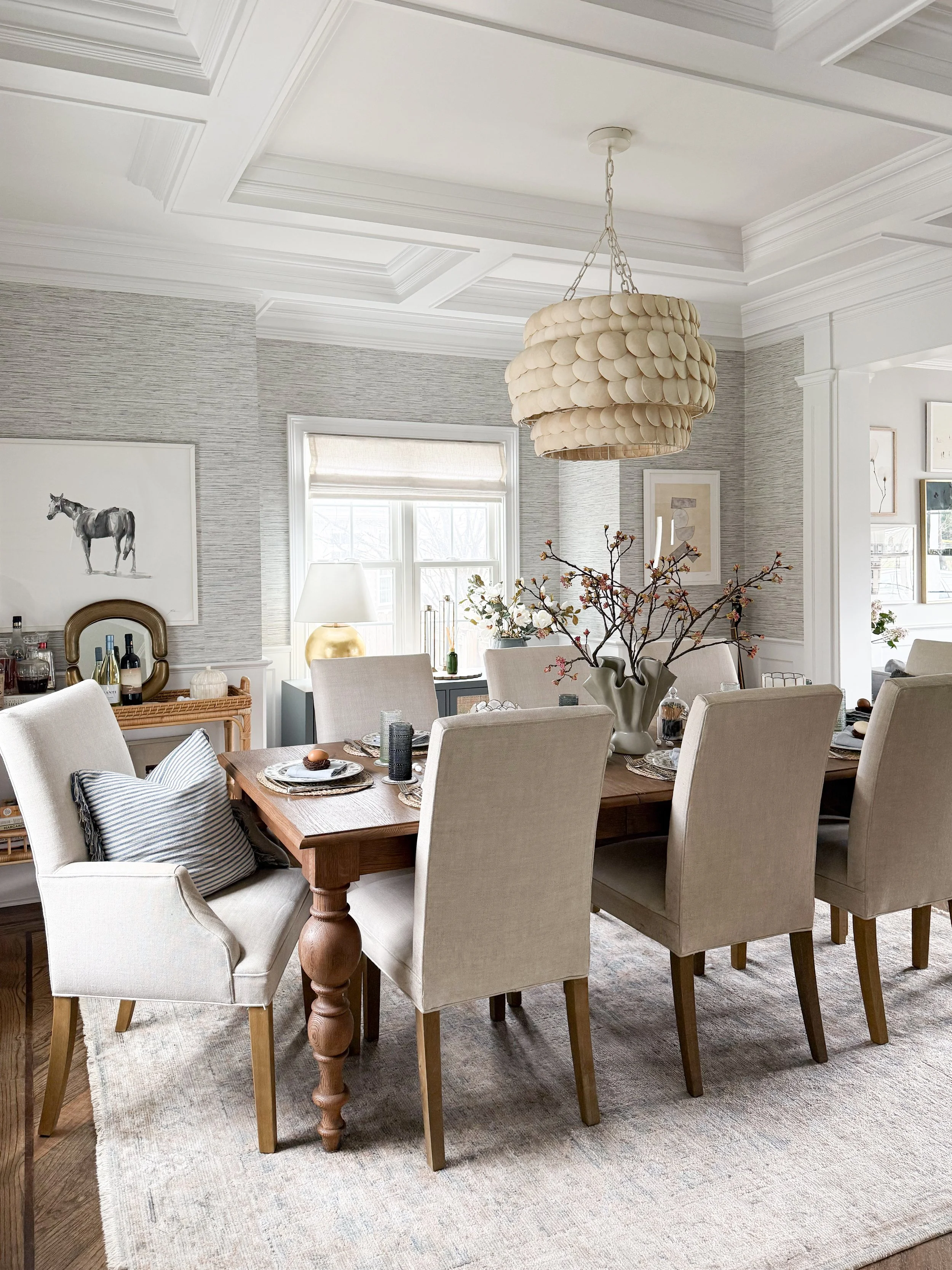 Neutral spring dining room with wood table, upholstered chairs, and elegant Easter tablescape featuring woven placemats and blooming branches centerpiece