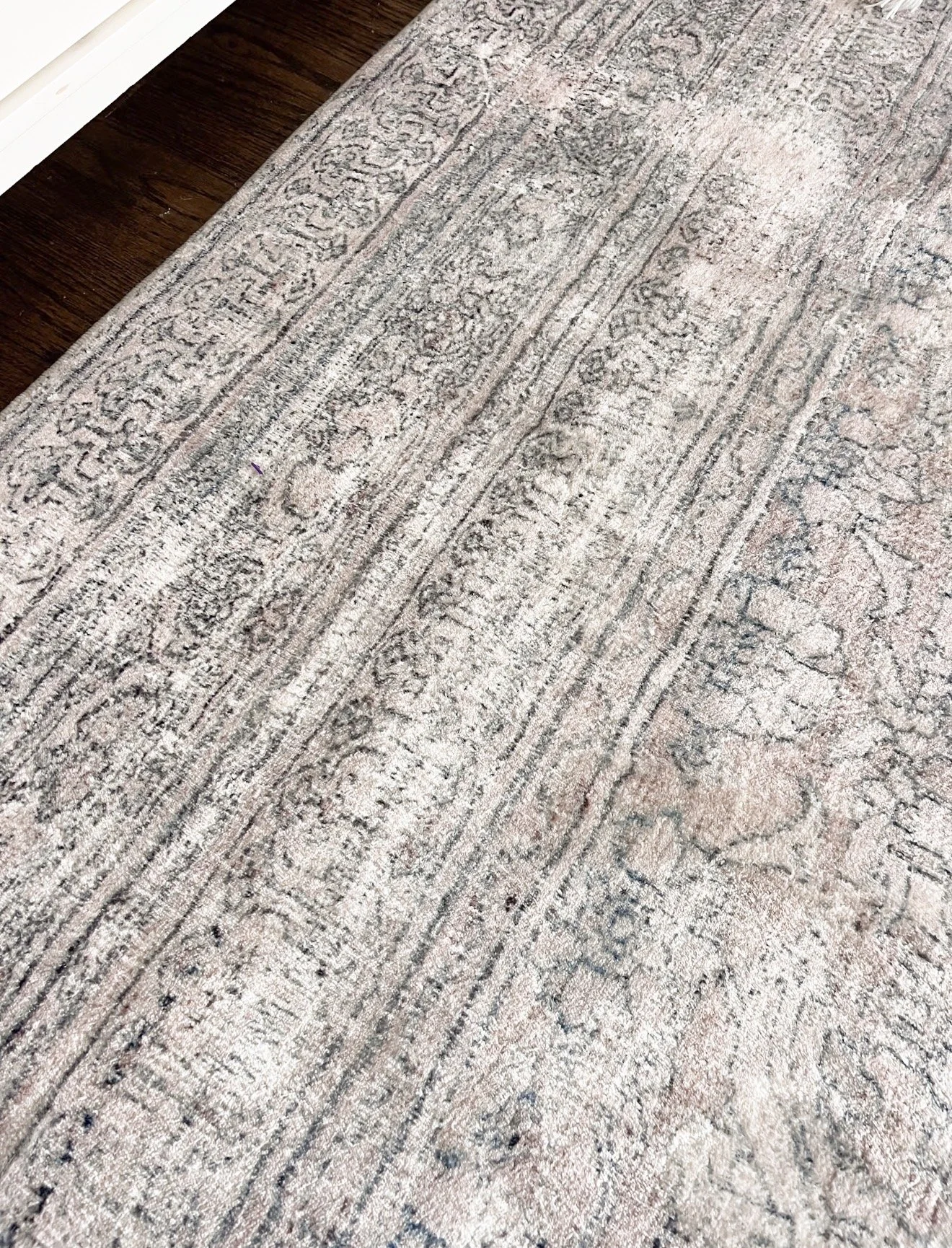 Close-up view of a muted blue and beige patterned area rug under the corner of a bed with white and gray bedding, showing the detailed floral and geometric design of the rug in a bedroom setting.
