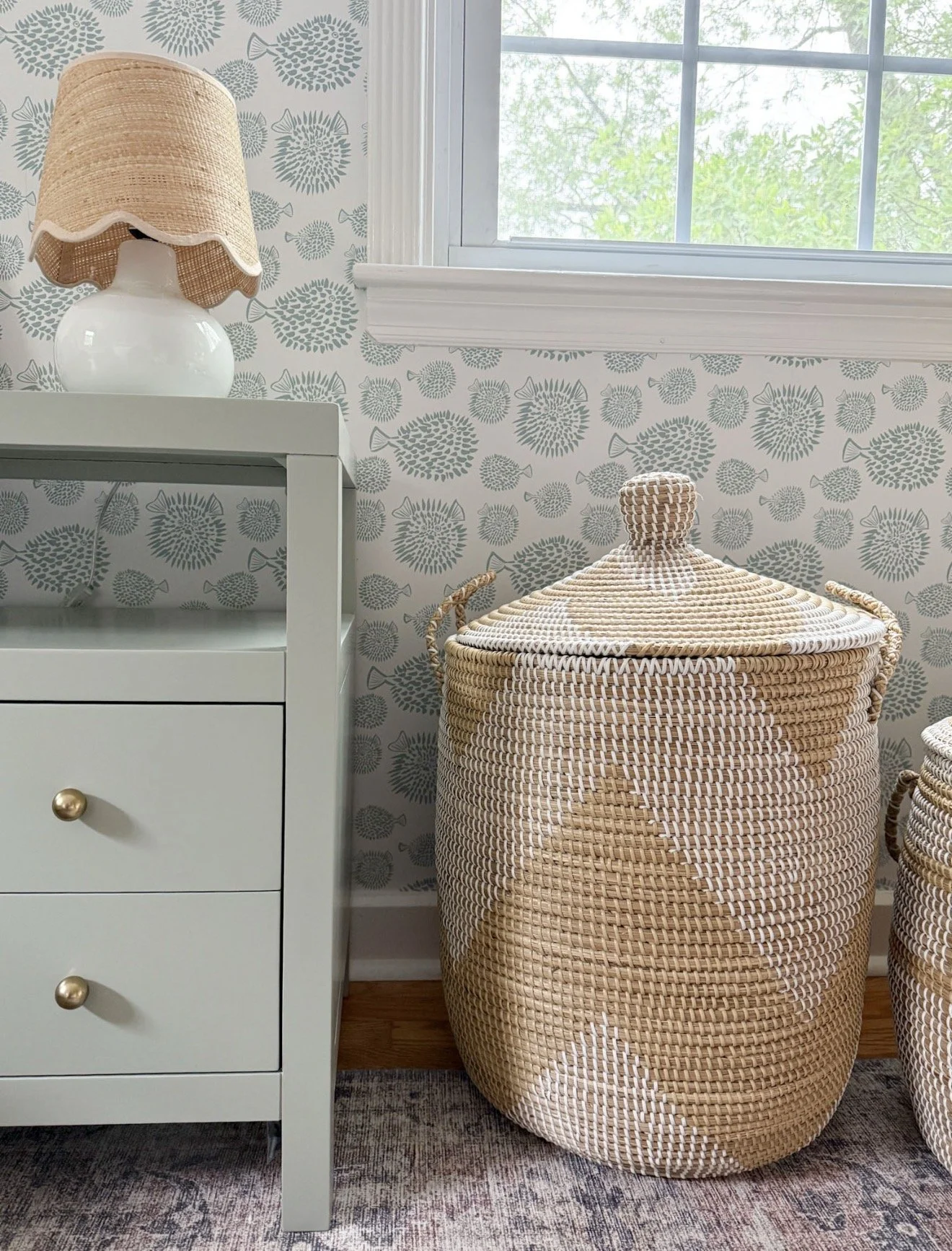 Close-up of soft blue patterned wallpaper paired with pink geometric curtains, a table lamp, and patterned pillow.