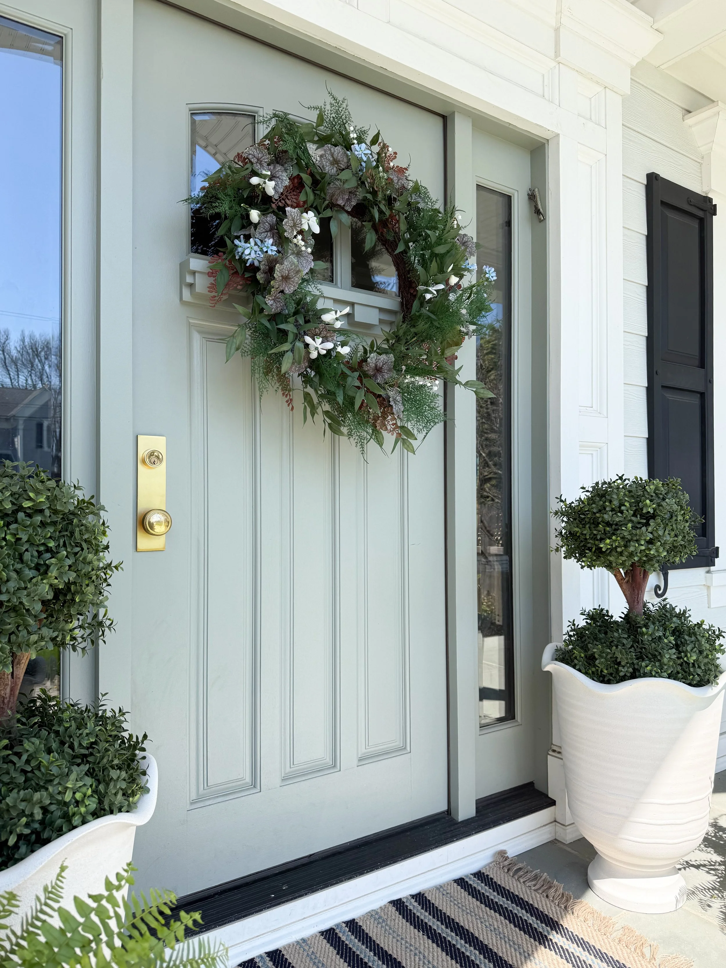 Symmetrical front porch styling with topiary planters, ferns, striped rug, and muted green door