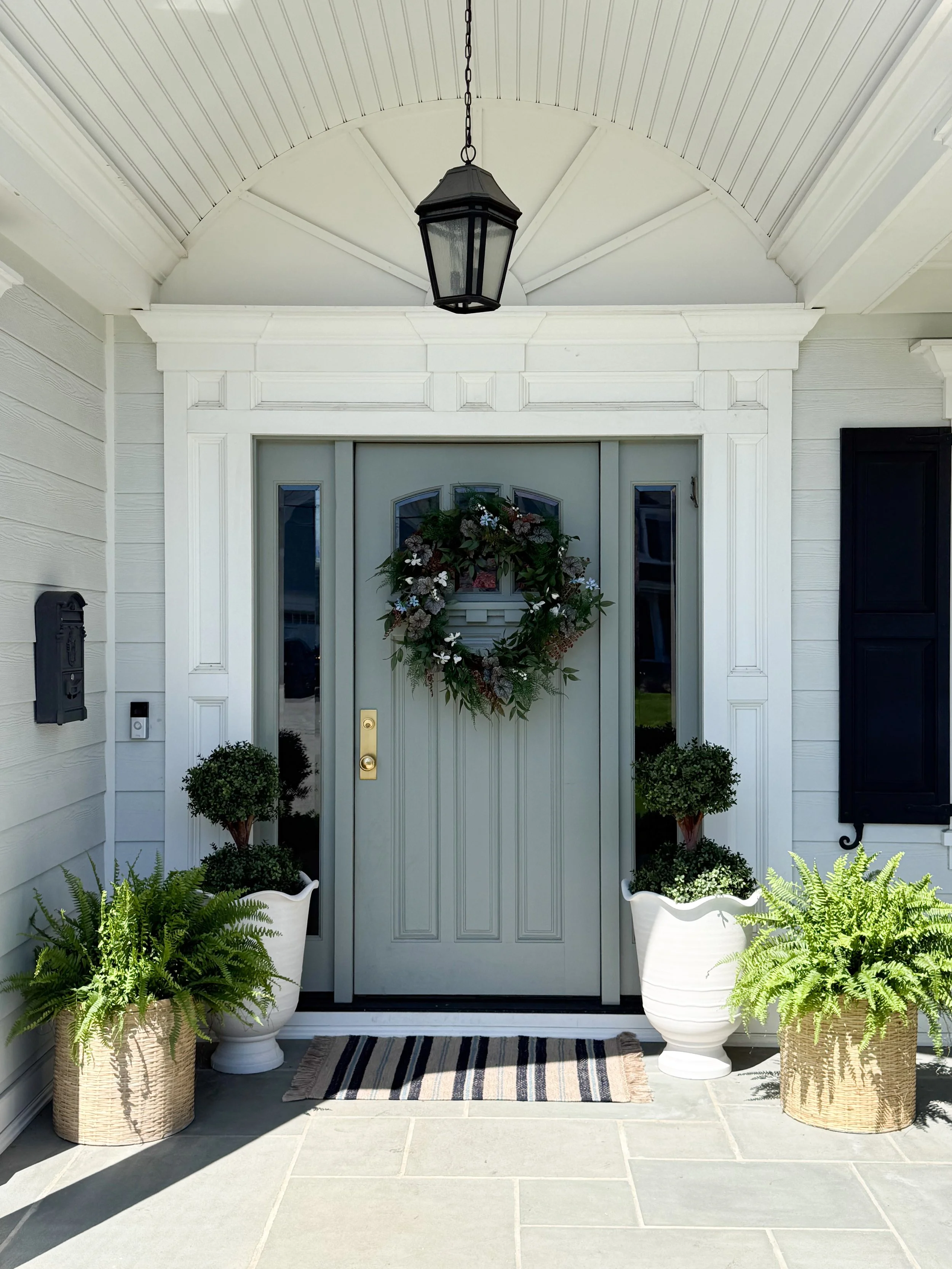 Spring front porch with muted green door, wreath, symmetrical planters, and striped welcome mat