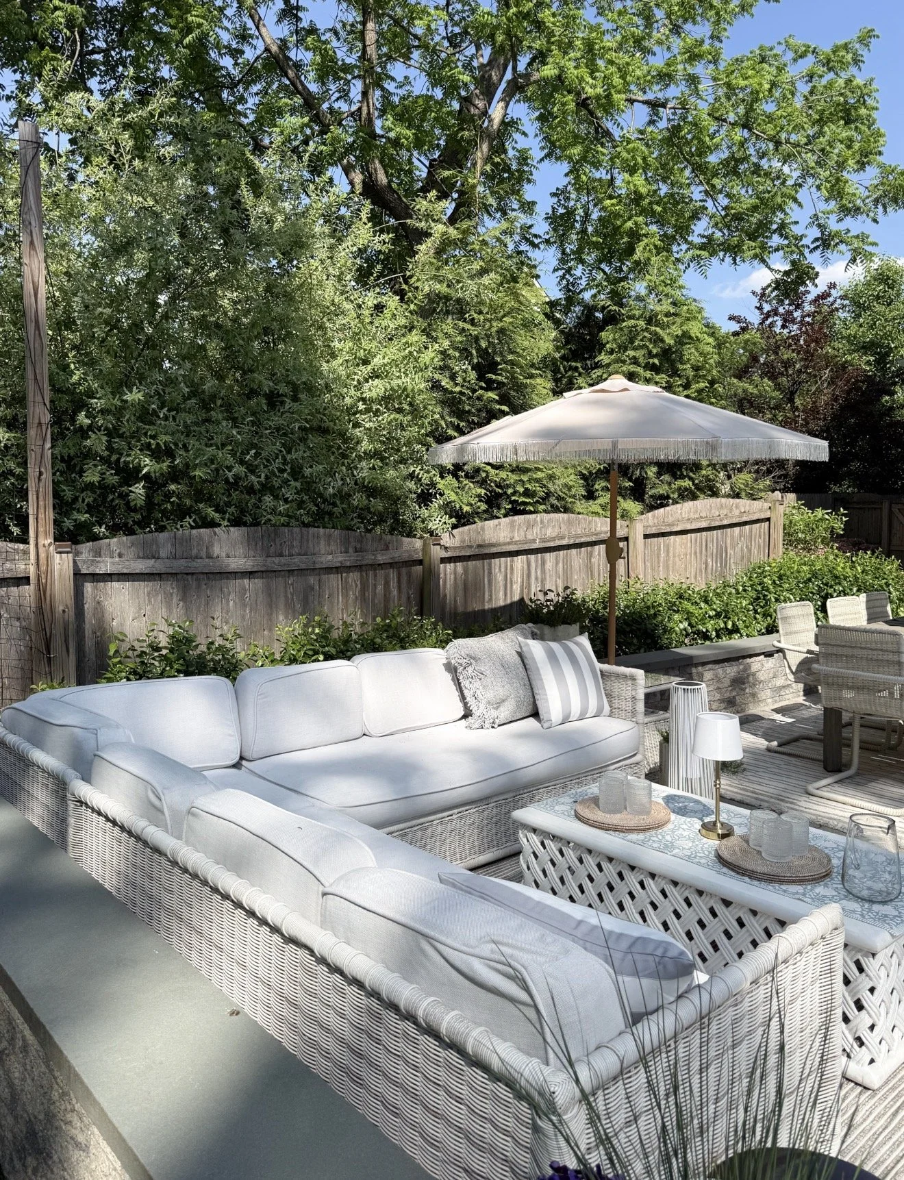 Neutral outdoor patio design with white woven lounge seating, layered coffee table styling, striped umbrella, and lush backyard landscaping surrounded by mature trees and greenery.