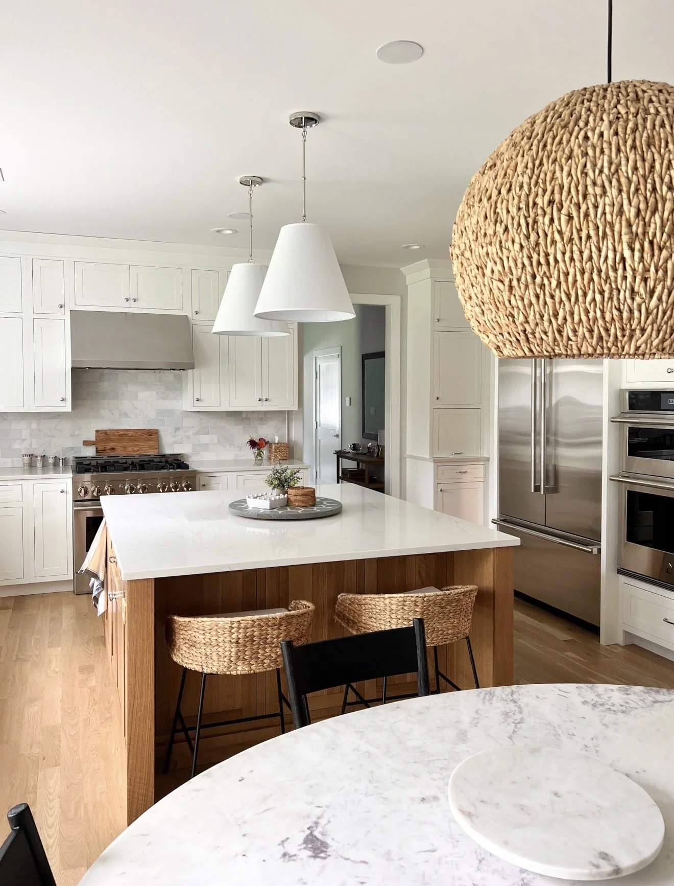 Bright kitchen with white pendant lights, woven statement pendant, large island, marble countertops, and woven counter stools.