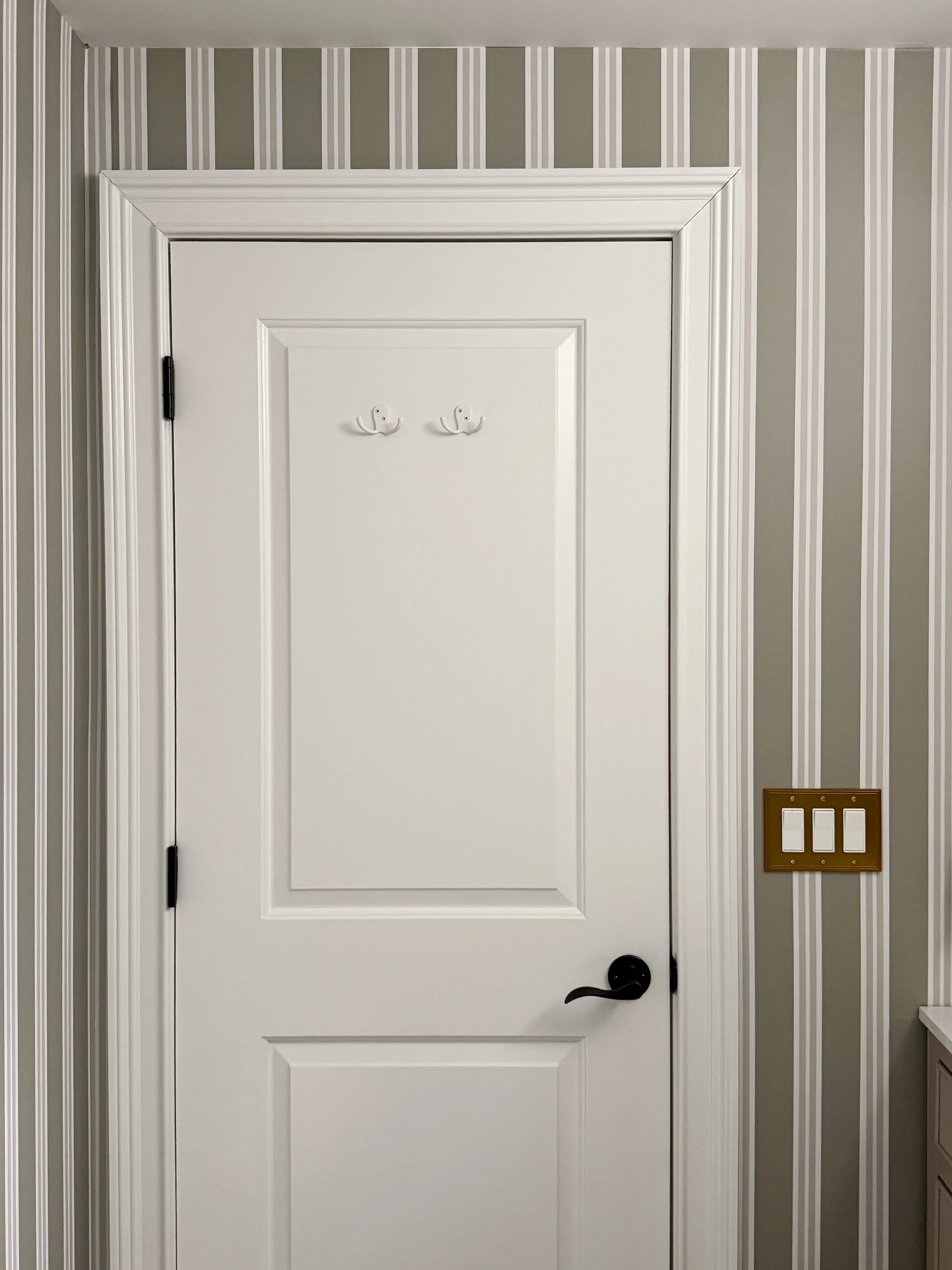 Basement bathroom door detail featuring Chasing Paper striped wallpaper, white paneled door, black hardware, and classic trim details in a neutral bathroom design.