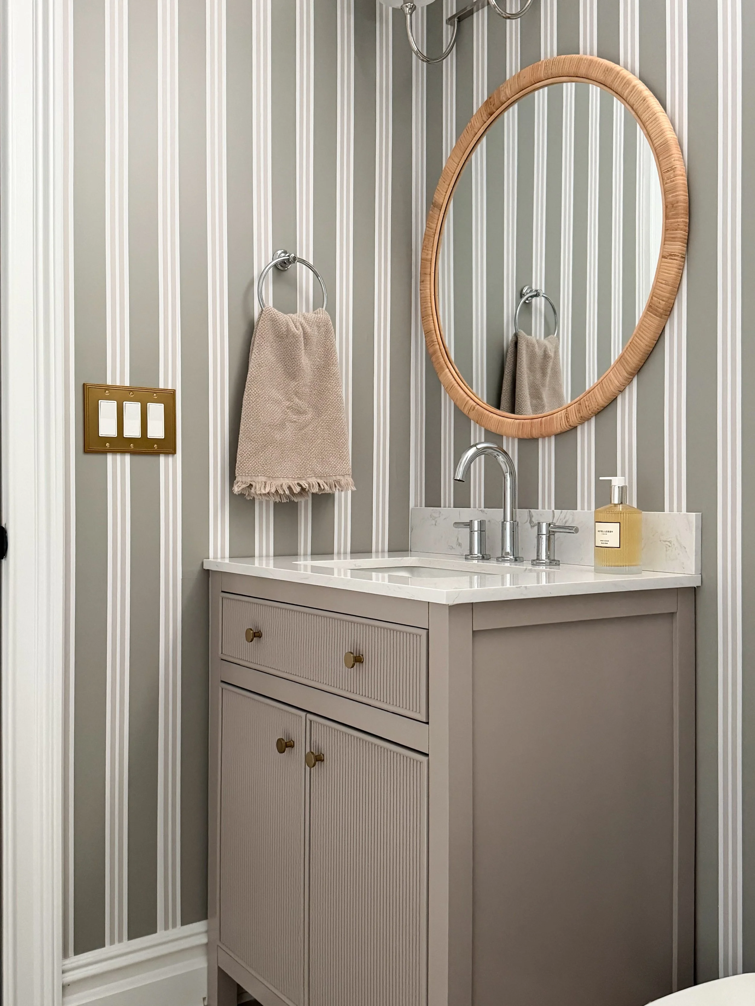 Small basement bathroom design featuring Chasing Paper striped wallpaper, a fluted vanity, rattan round mirror, marble countertop, chrome faucet, and neutral decor accents.