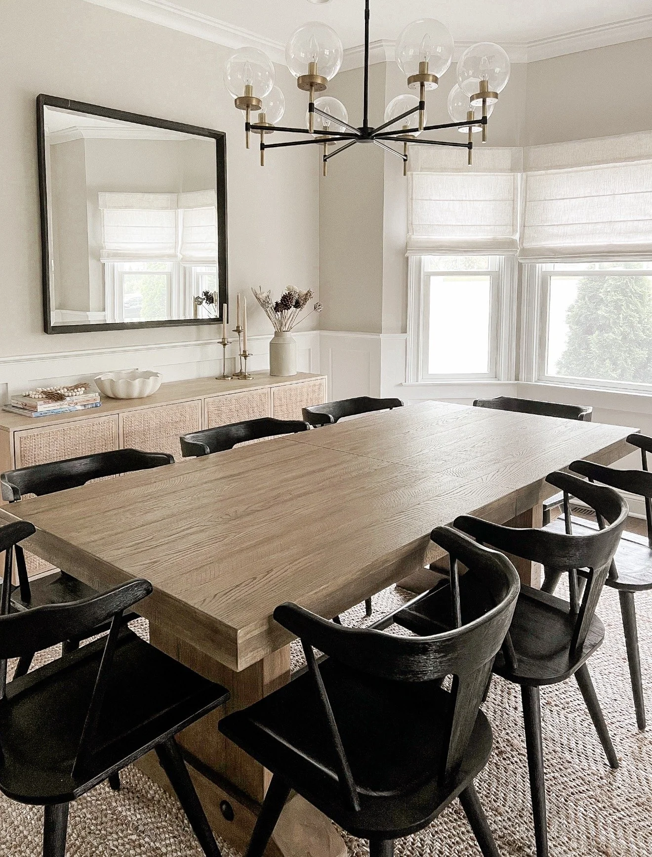 Modern dining room with black and brass chandelier, large wood dining table, black dining chairs, and woven sideboard.