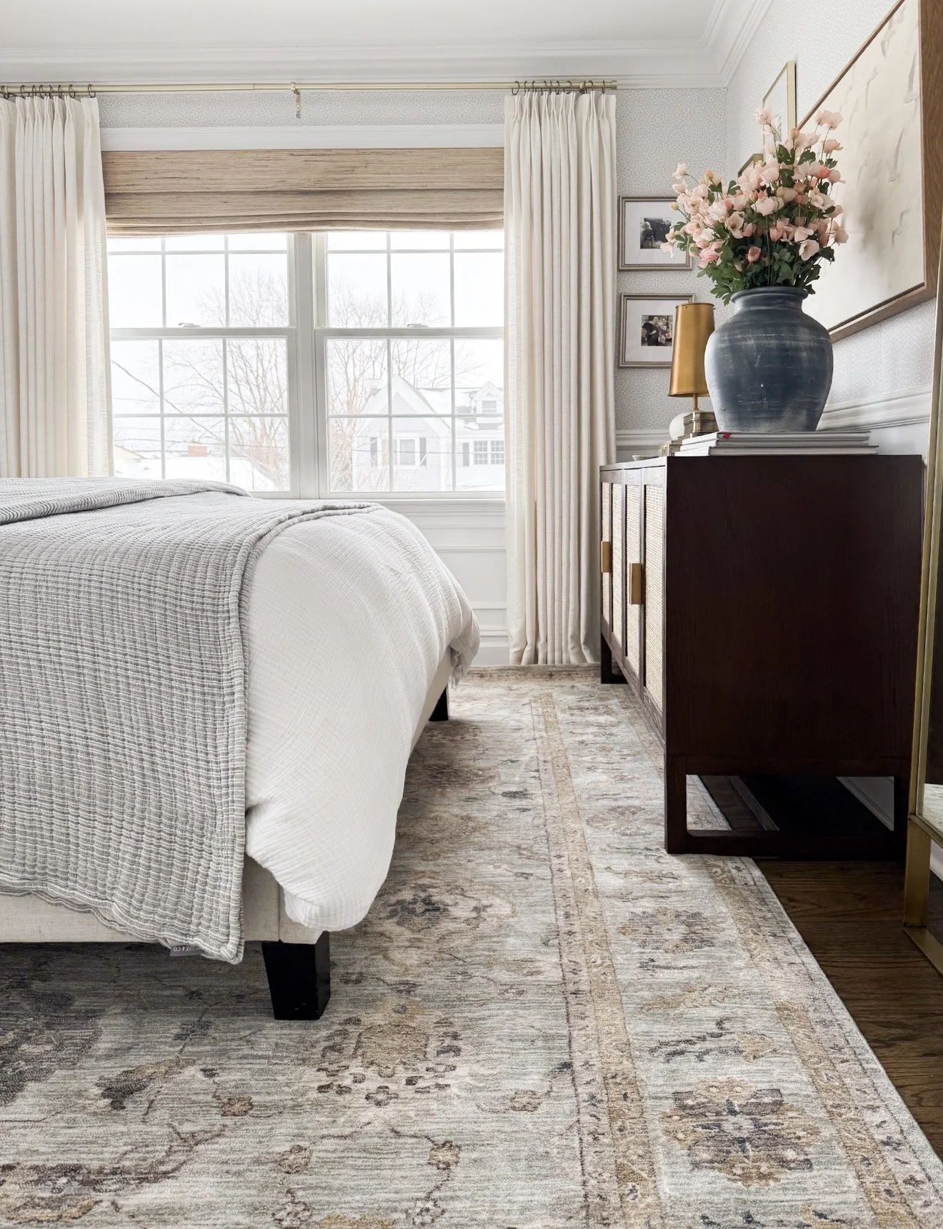 Soft neutral Loloi area rug layered beneath a bed with white bedding, a dark wood dresser, cream curtains, and a blue vase filled with pink flowers.