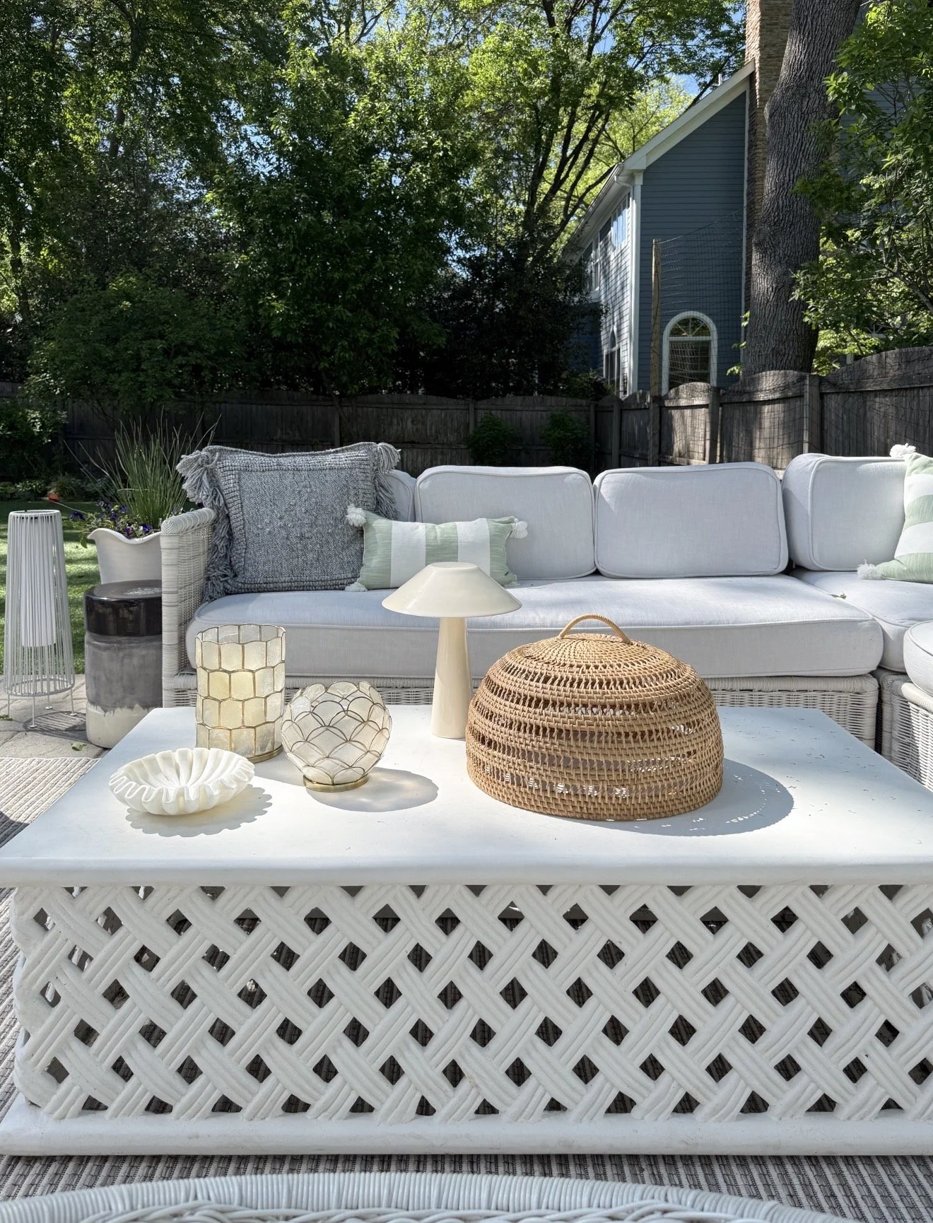 Coastal inspired outdoor patio with white woven furniture, lattice coffee table styling, neutral decor accents, and layered backyard landscaping surrounded by mature trees.