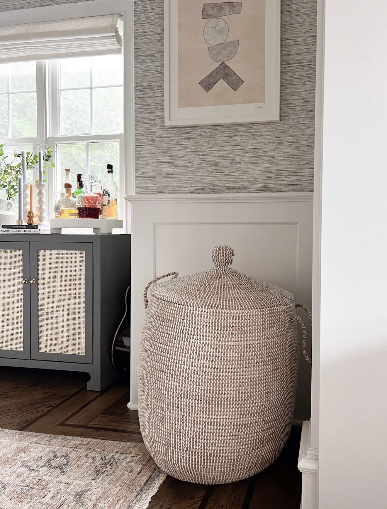 Round woven Serena and Lily La Jolla basket in beige with subtle white accents placed next to a dark blue bar cabinet with cane doors in a living room.
