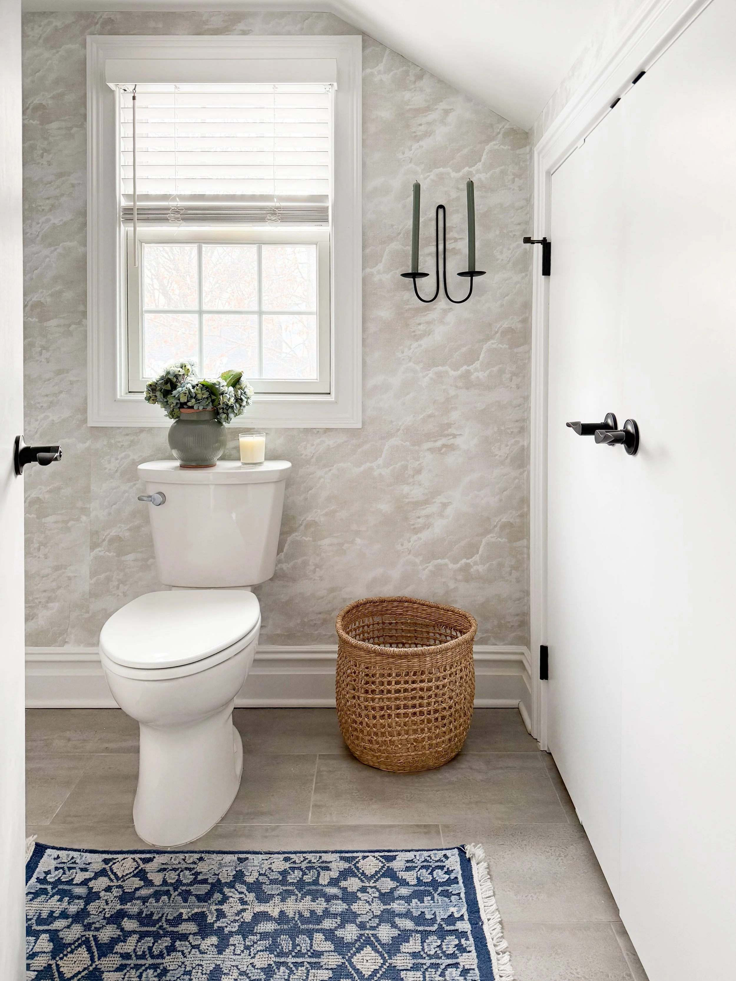Bright bathroom with a white toilet beneath a window with blinds, styled with a vase of hydrangeas and a candle, textured neutral wallpaper, a black double candle sconce, woven basket, and blue patterned rug on tile flooring.