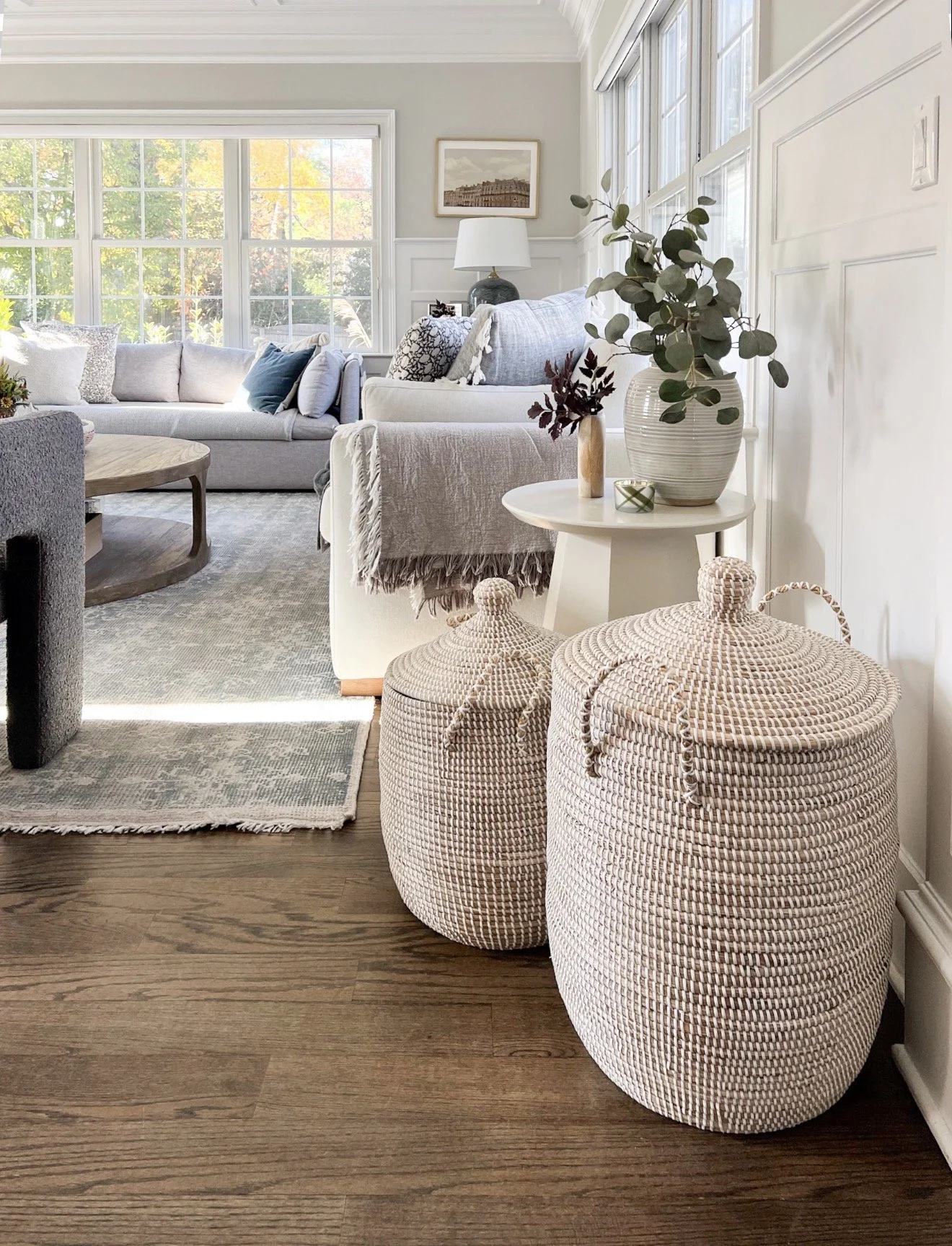 Two large woven round storage baskets with lids placed on a hardwood floor next to a white side table, in a bright living room with a light gray rug, soft sofas, and natural sunlight coming through the windows.