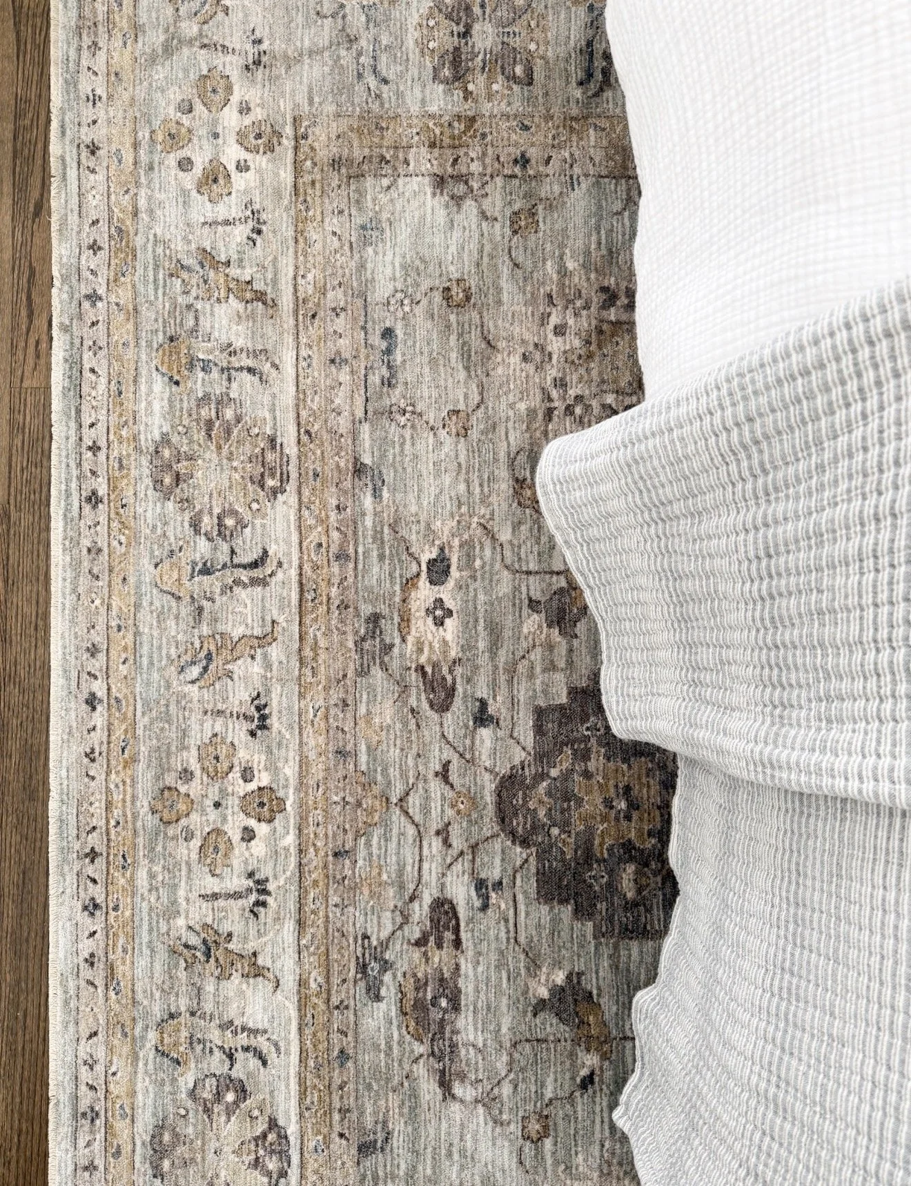 Close-up of a vintage-inspired Loloi rug with muted blue, taupe, cream, and brown detailing beside layered white and gray bedding.