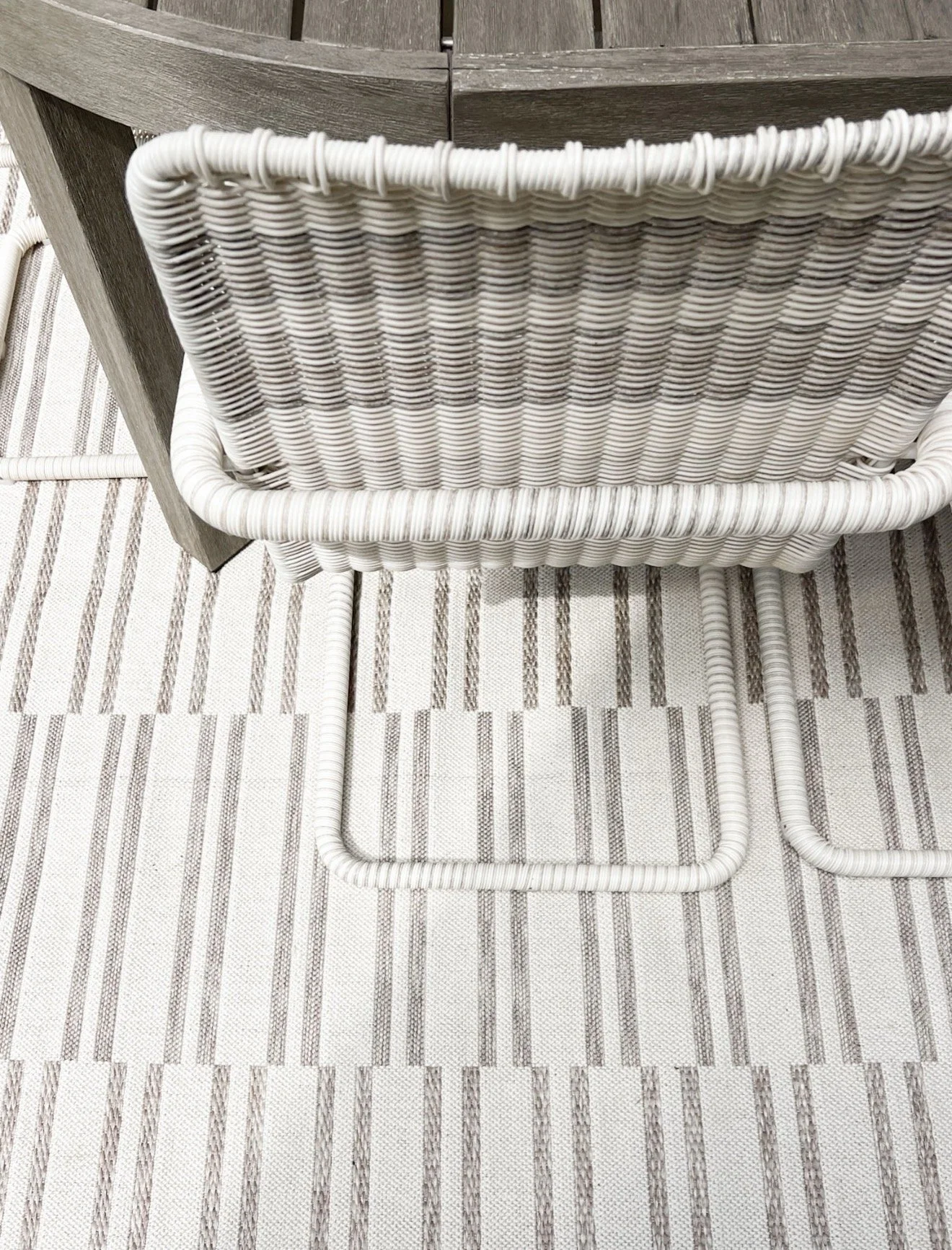 Neutral coastal patio seating area with wicker sectional sofa, gray outdoor pillows, woven coffee table, and striped outdoor rug.