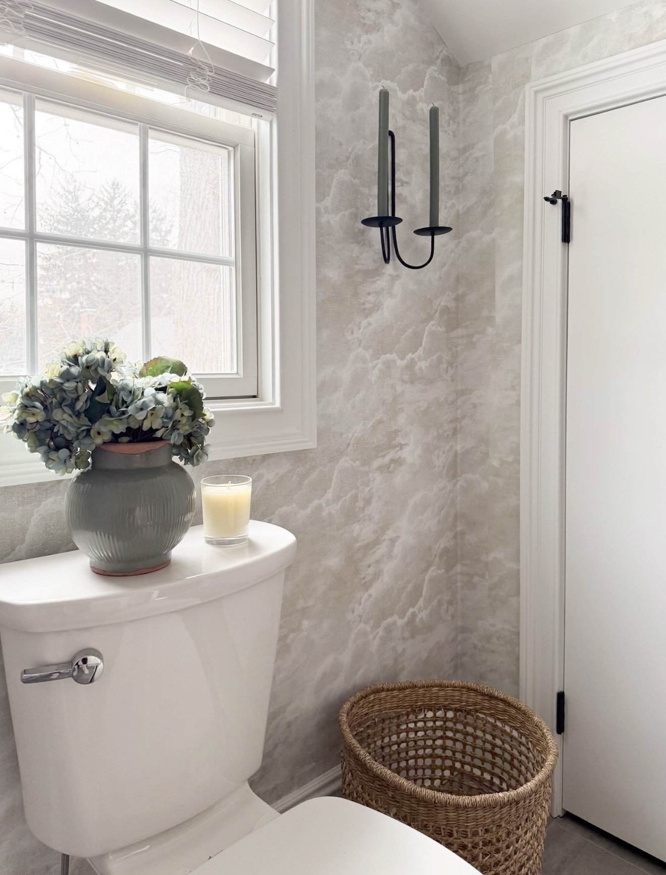 Neutral powder bathroom with black taper wall candle holder, soft textured wallpaper, hydrangea arrangement, woven basket, and elevated traditional styling