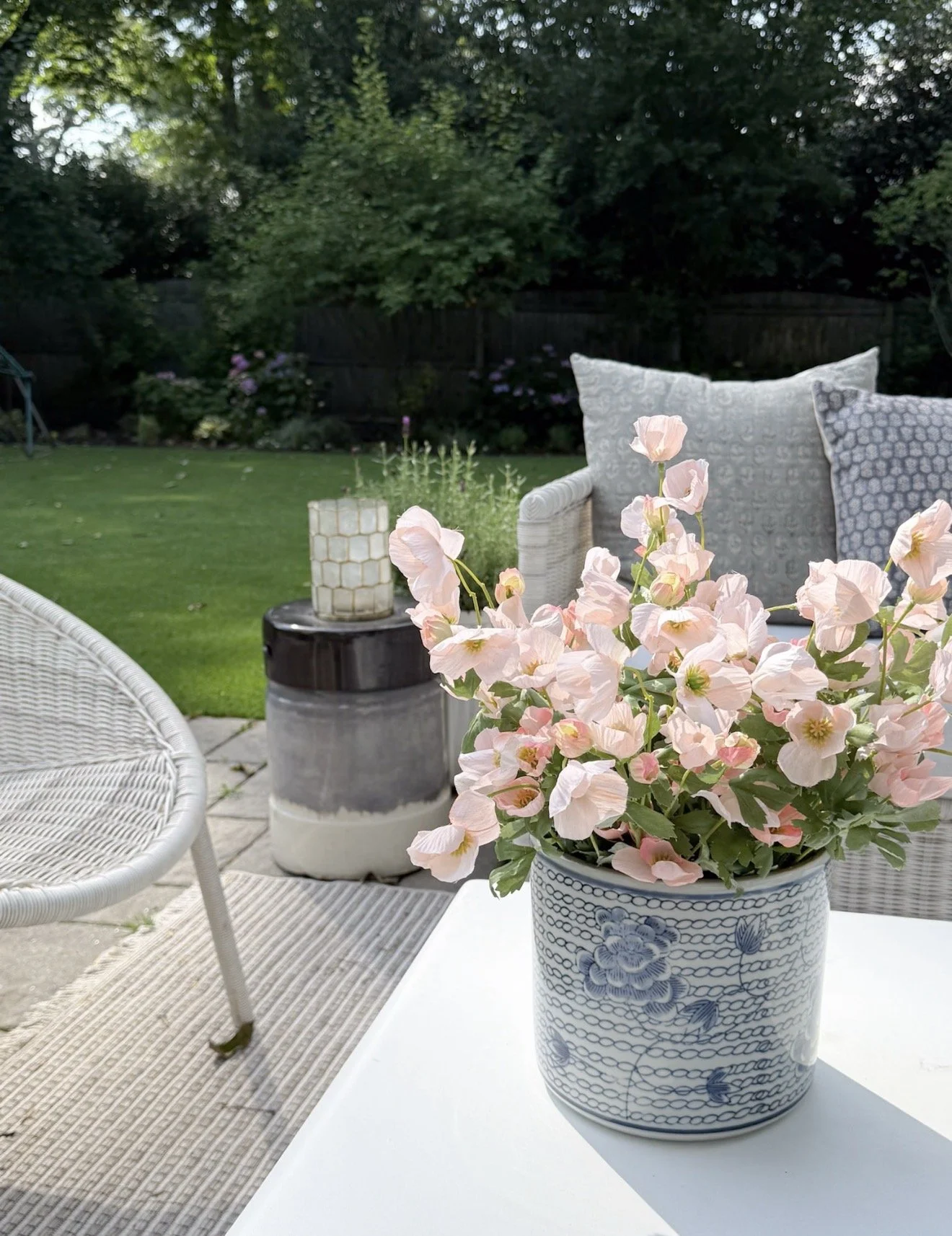 Coastal outdoor coffee table styling with woven white table, blue glass candlesticks, decorative candle holders, and pink flowers in a blue planter.