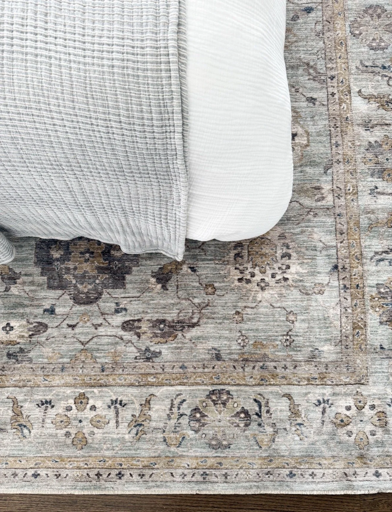 Overhead view of a bedroom rug in muted gray and beige tones with traditional patterns, partially covered by a bed with white sheets and a light gray striped blanket