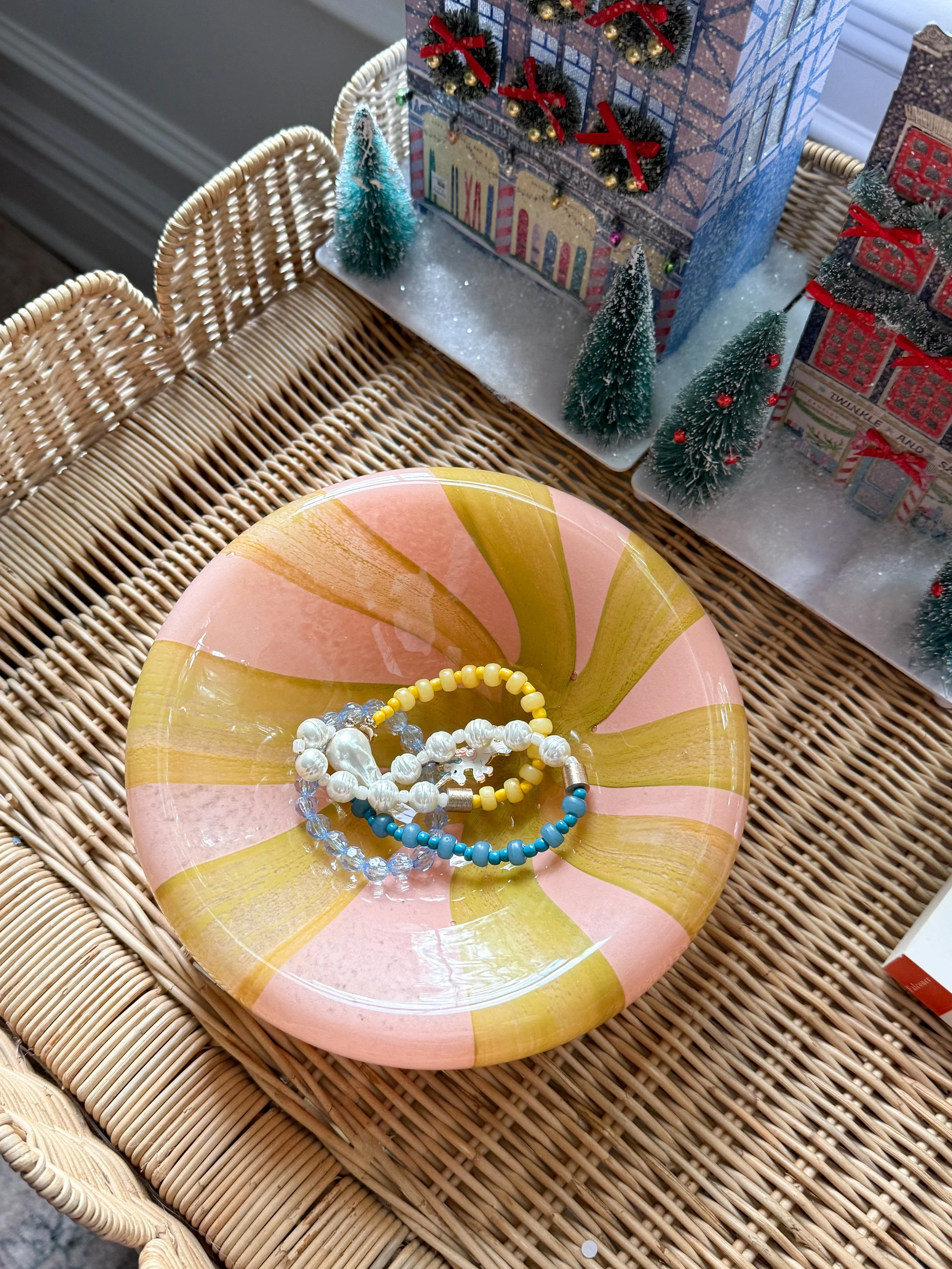 Anthropologie holiday decor featuring a pink and yellow striped decorative dish with colorful beaded bracelets, styled beside miniature Christmas village houses on a woven rattan table.