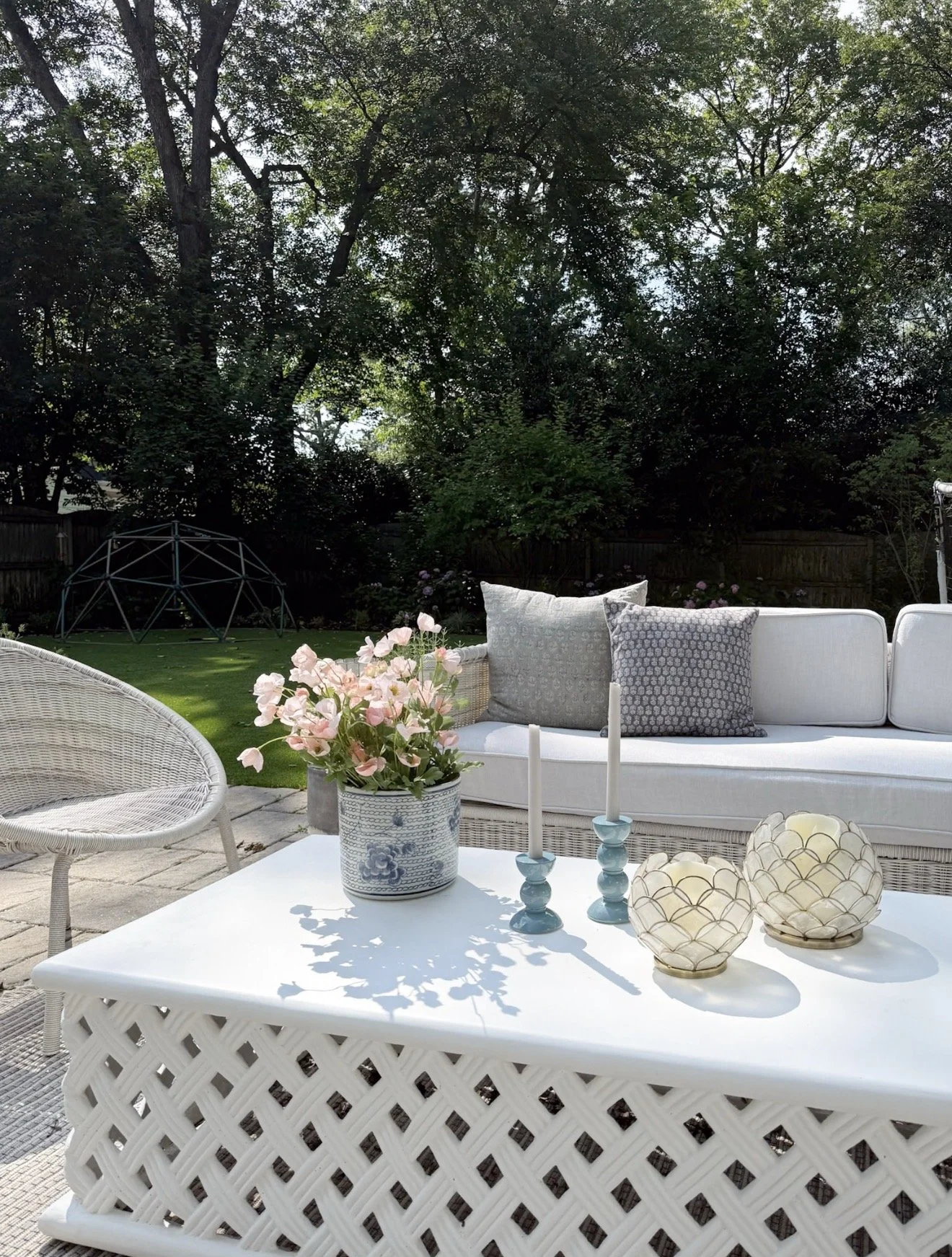 Coastal outdoor coffee table styling with woven white table, blue glass candlesticks, decorative candle holders, and pink flowers in a blue planter.