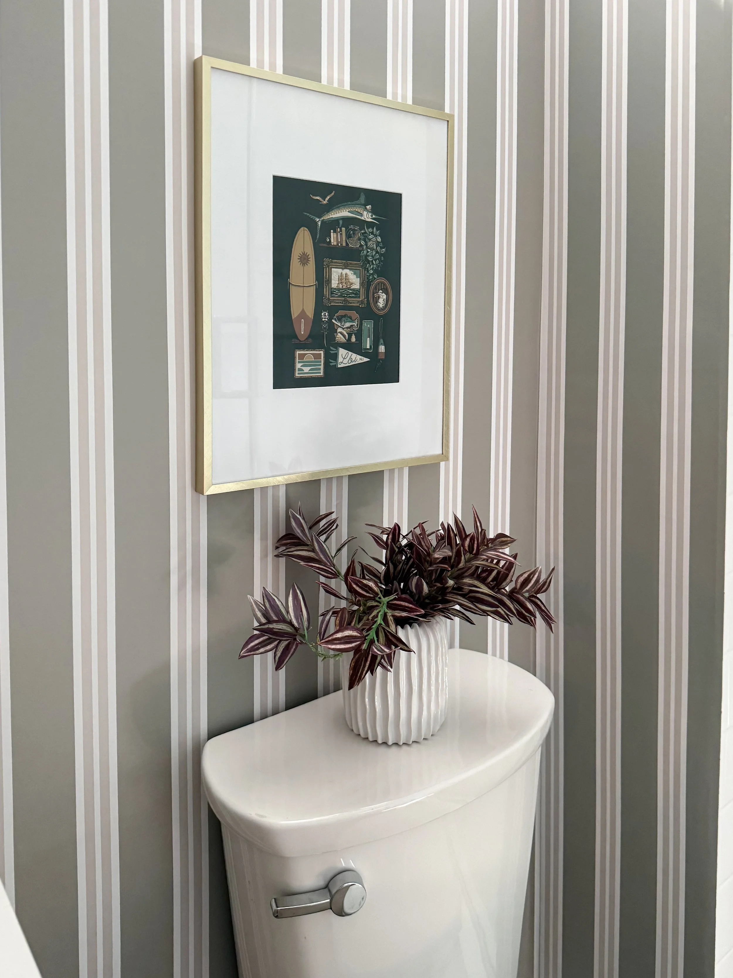 Basement bathroom styling detail featuring Chasing Paper striped wallpaper, framed artwork, a textured white vase with greenery, and a neutral toilet area design.