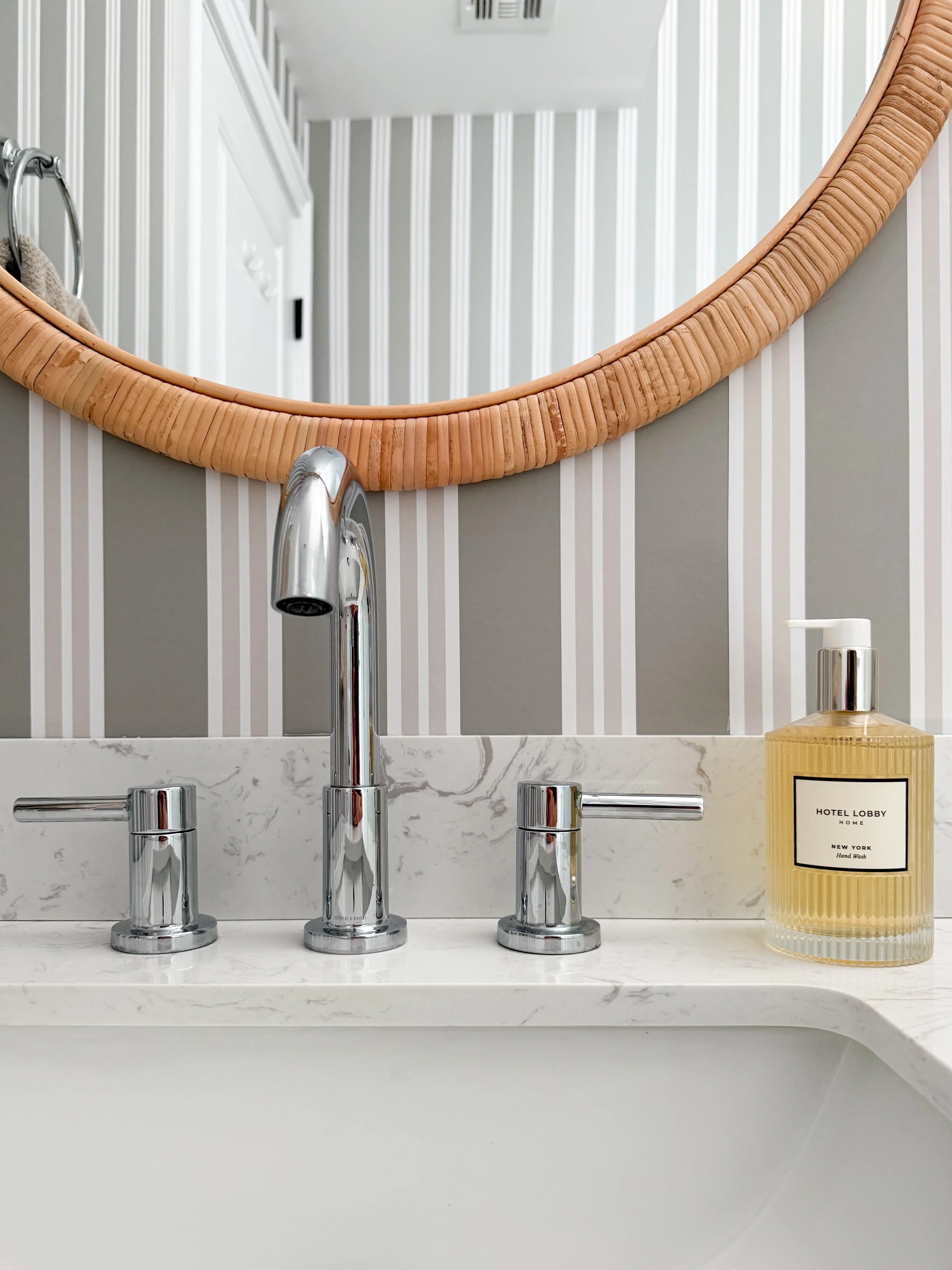 Bathroom vanity detail featuring a rattan round mirror, chrome faucet fixtures, marble countertop, striped Chasing Paper wallpaper, and neutral sink styling.