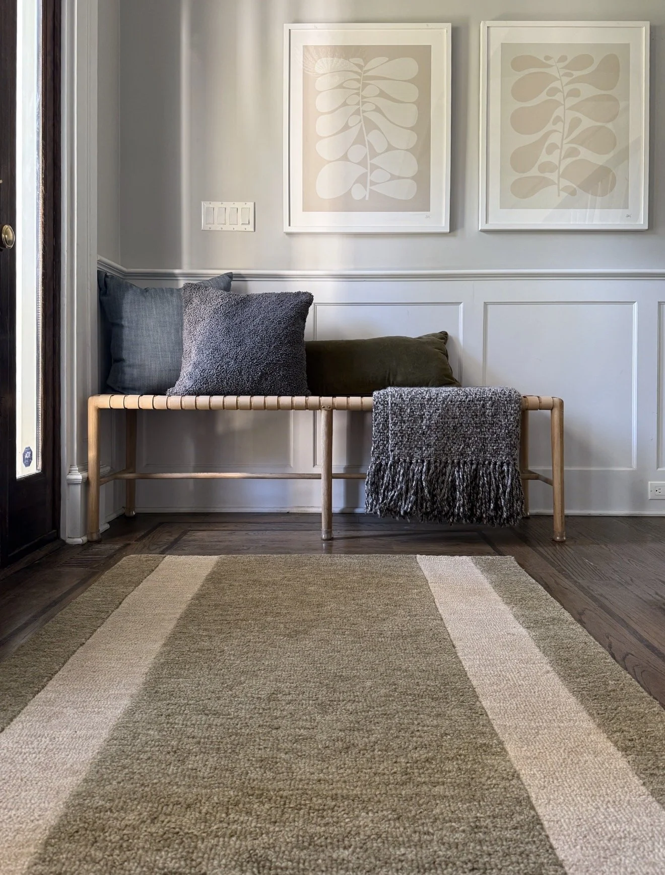 Entryway with a woven bench, layered neutral pillows, framed botanical art, and a striped green and cream Loloi area rug.