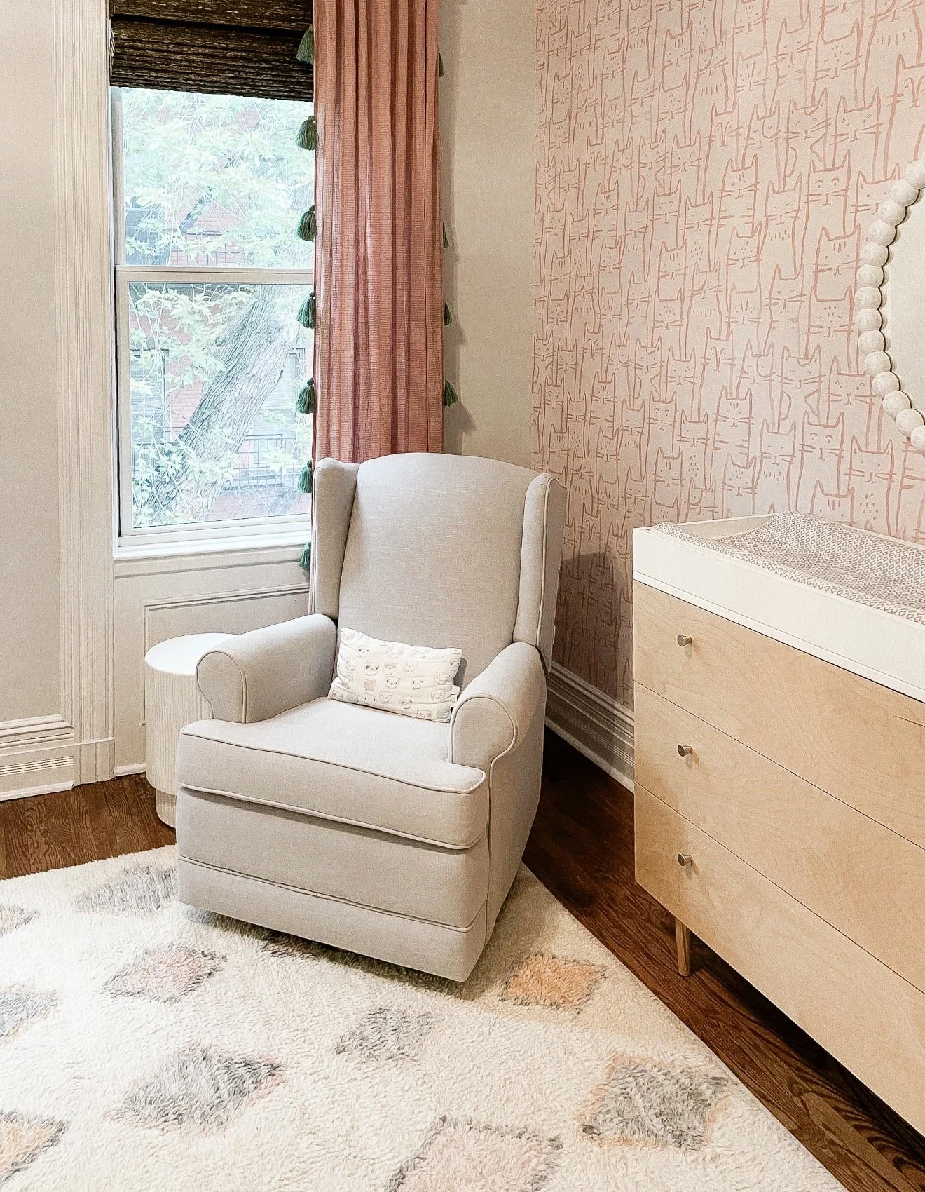 Nursery with pink cat wallpaper, pink curtain panels, neutral glider, wood dresser, and patterned area rug.