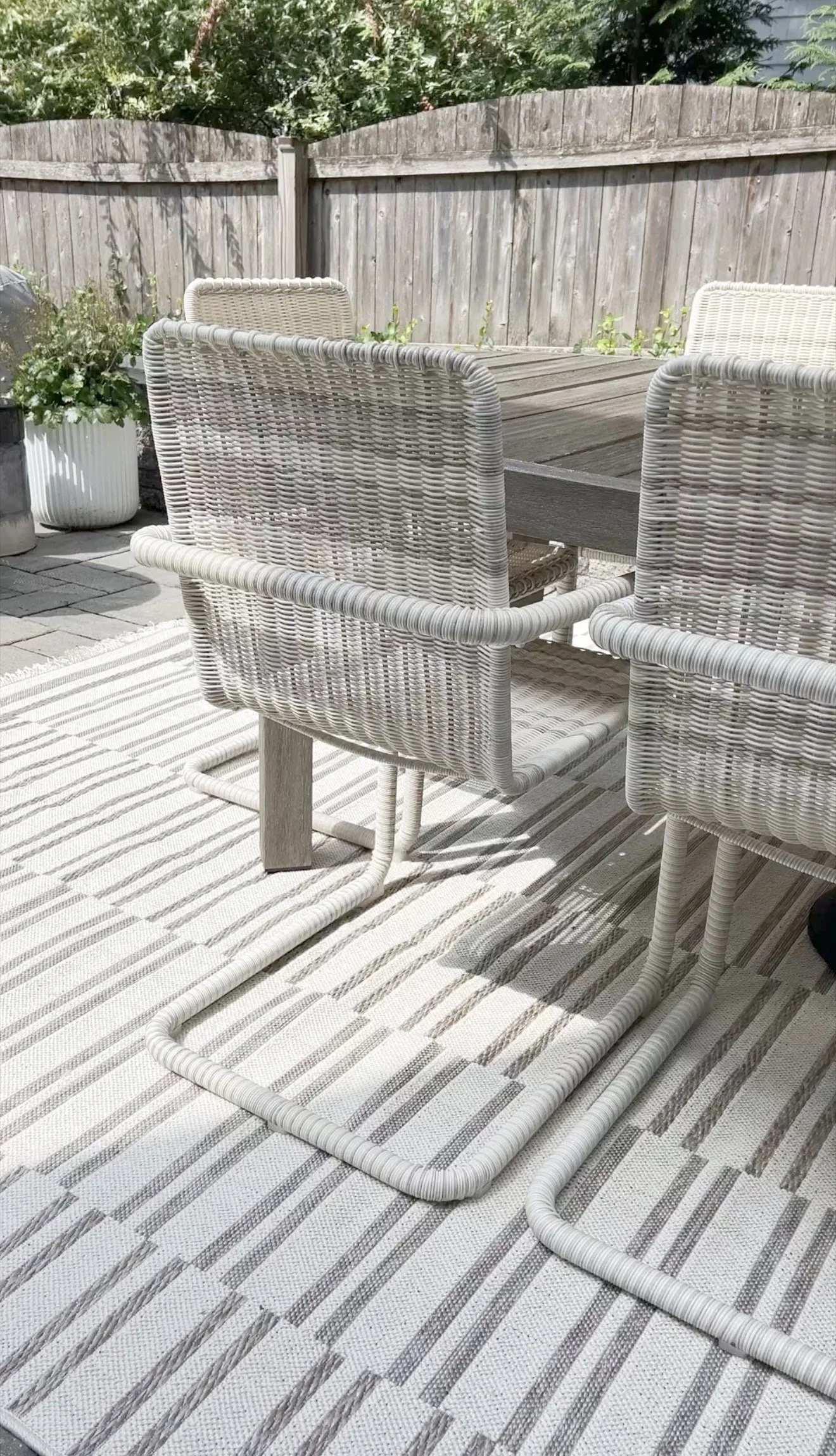 Outdoor patio scene showing the back of two white woven chairs positioned around a gray wooden dining table.