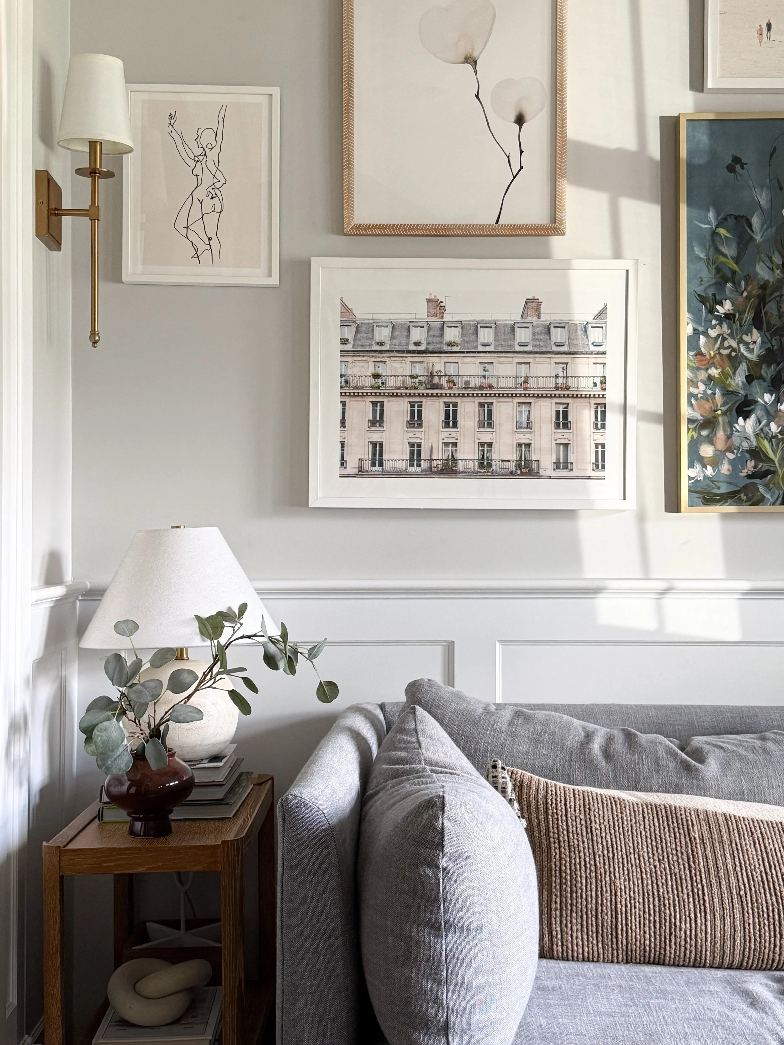 Neutral living room styled for fall with a gray sofa, warm brown textured pillow, eucalyptus branches, layered artwork, and soft natural light.