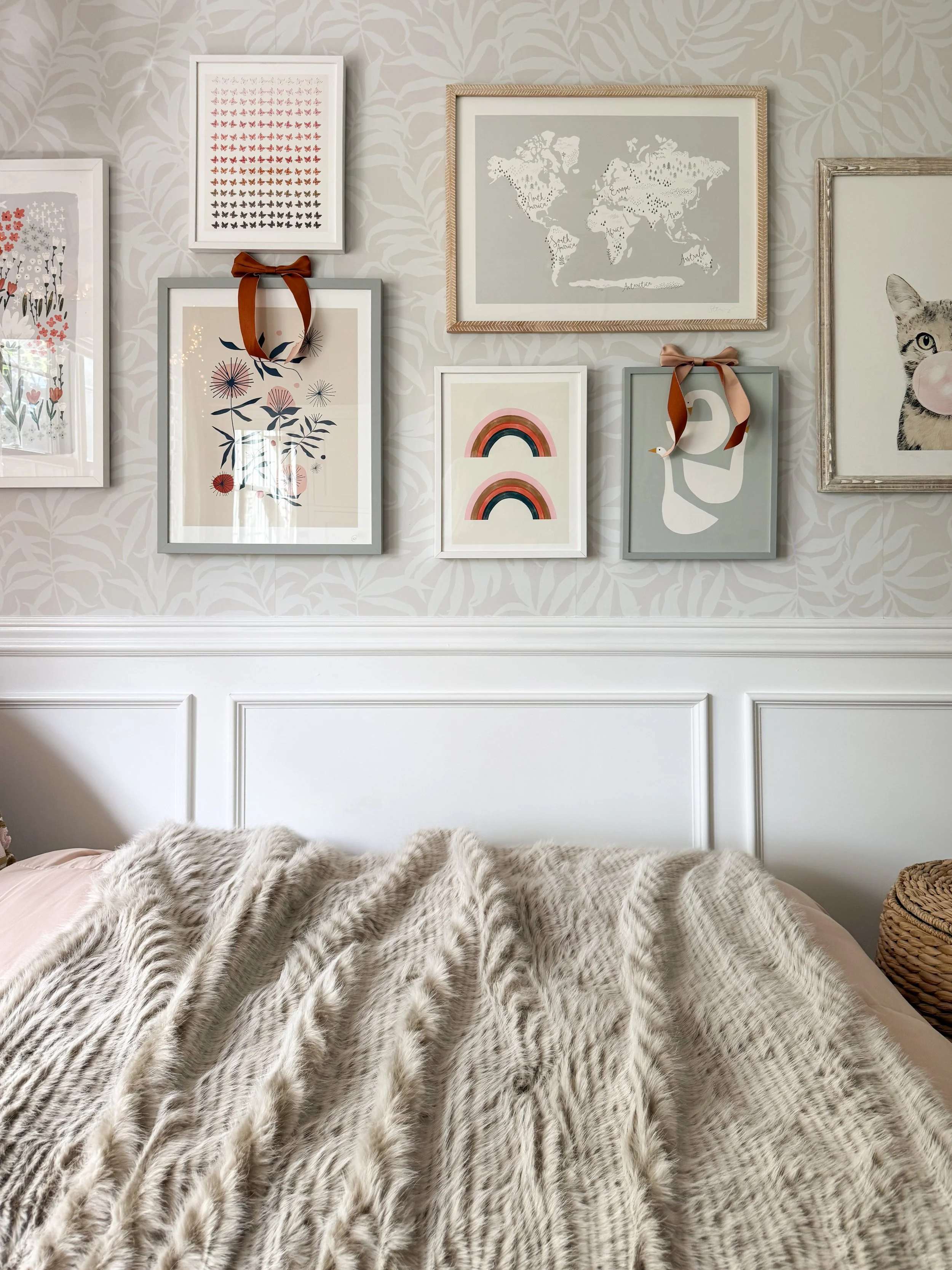 Anthropologie holiday bedroom decor featuring a gallery wall of colorful framed artwork with ribbon accents above a bed styled with a plush faux fur throw and pale pink bedding.