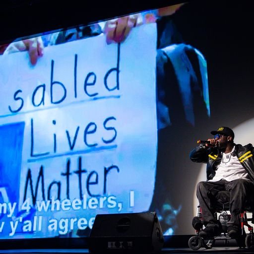 Image of a male performer in a wheelchair singing on stage in front of a video screen with the image of a disabled lives matter sign at The Danny Awards.
