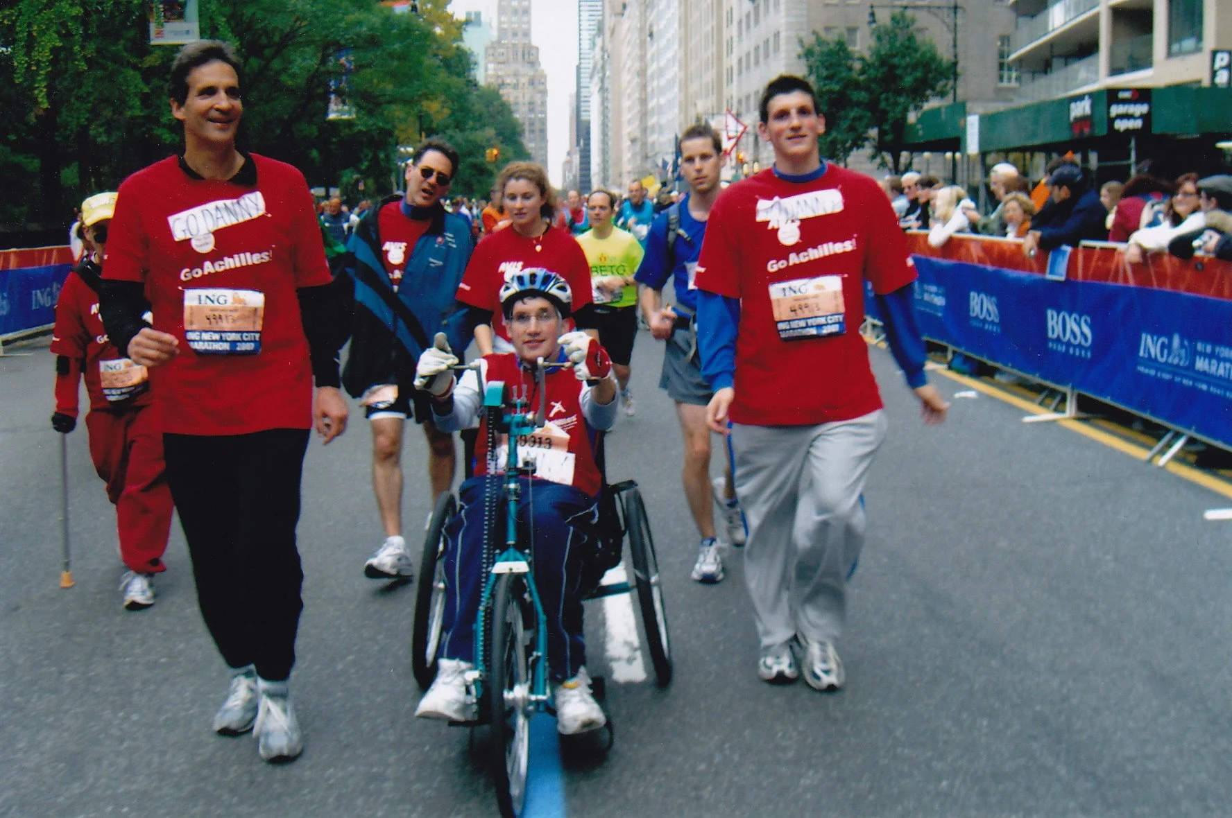  Image of Daniel and his family and guides at the NYC Marathon. Daniel is riding a hand cycle, while his father, brother and guides walk alongside him. 