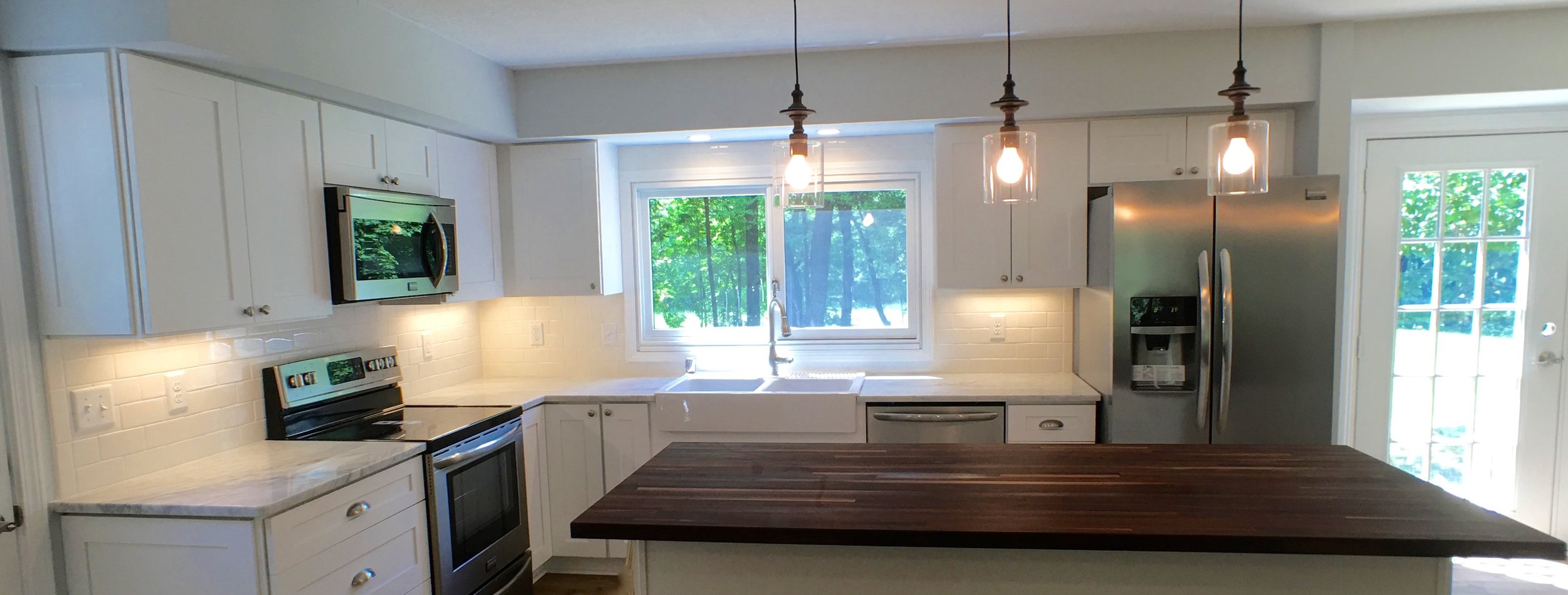 White kitchen with marble countertops, walnut butcher block, pendant lighting, stainless steel appliances, white farmhouse sink, subway tile backsplash, and under cabinet lighting