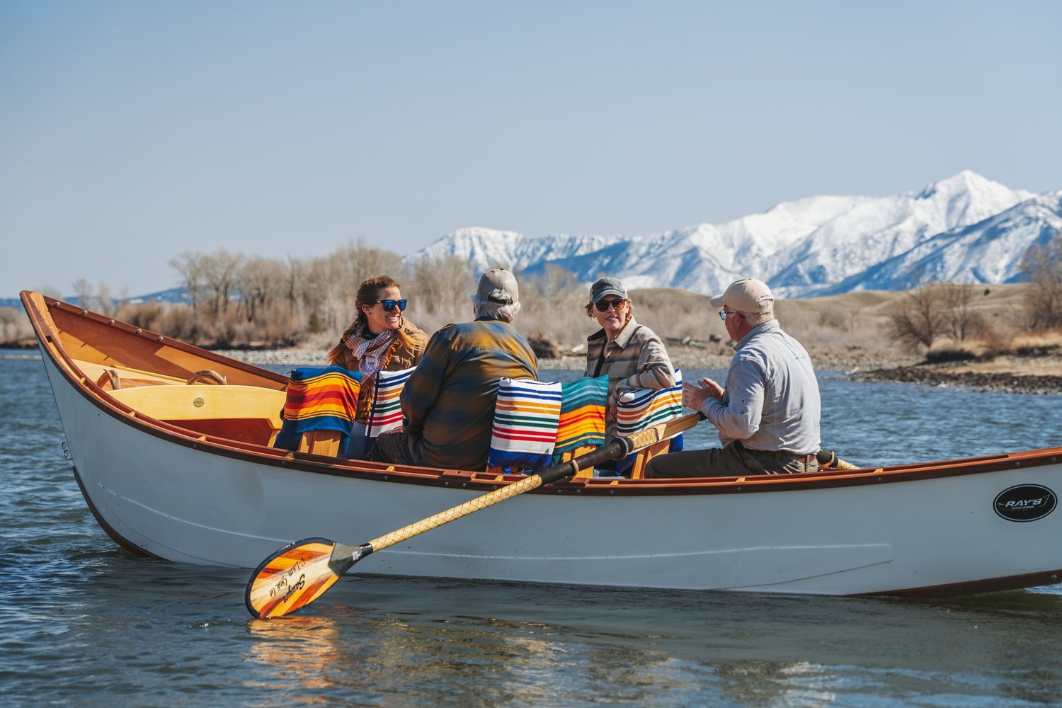 Guests enjoying a scenic wooden boat float on the Yellowstone River in a handcrafted Ray's River Dory with mountain views and Pendleton blankets near Livingston, Montana.