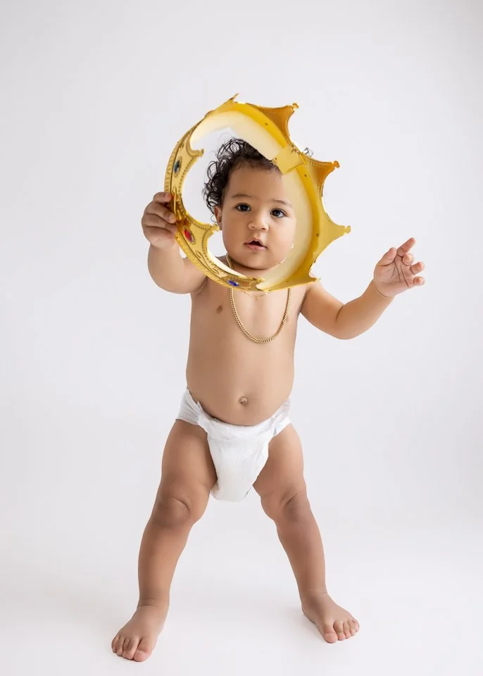 A toddler wearing only a diaper and a gold chain, holding a partially deflated yellow balloon and looking at the camera, against a plain white background.