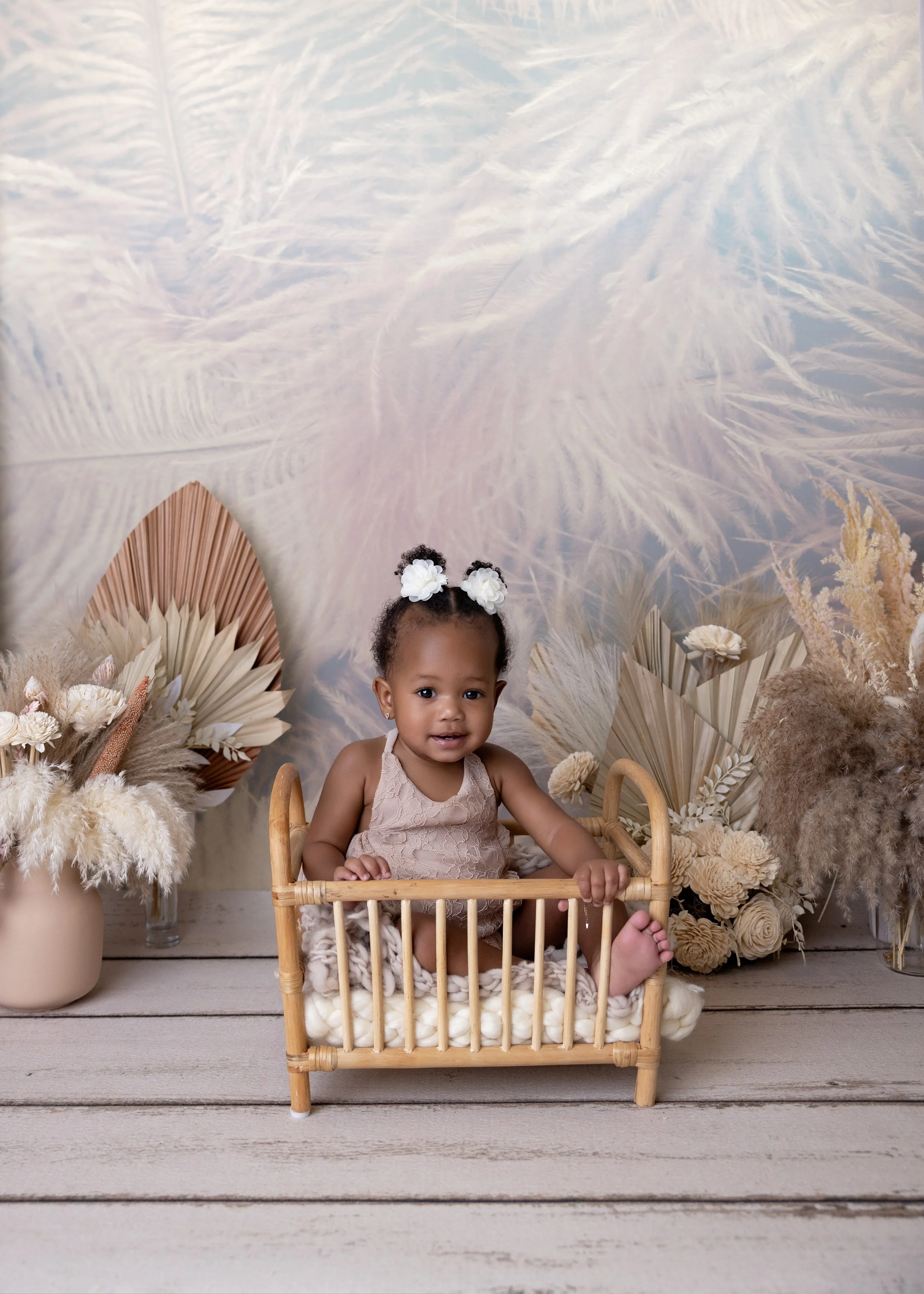 A young girl with curly hair and a white flower headband sitting in a small wooden bed, surrounded by beige and white dried floral arrangements and a pastel feathered patterned backdrop.