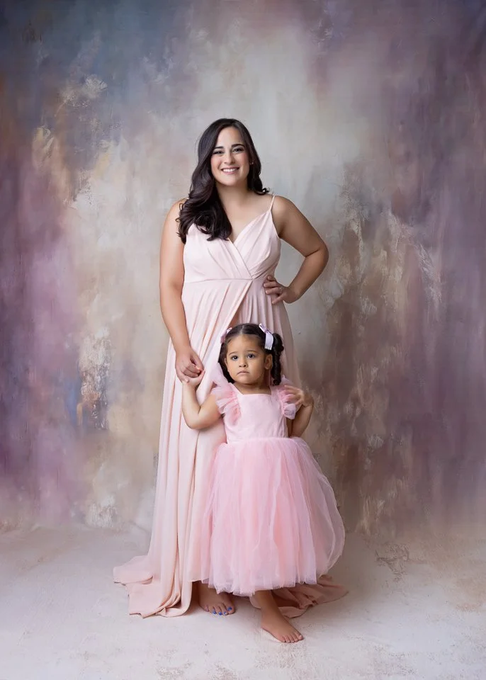 A woman and a young girl standing together against a painterly background. The woman is in a long, flowing pale pink dress, smiling, with her hand on her hip. The girl, also in a pink dress, looks slightly serious, holding the woman's hand, with a bo