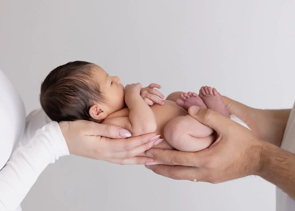 A newborn baby lying on its back being cradled by two adults' hands, one adult supporting the baby under its head and shoulders, the other holding its legs.