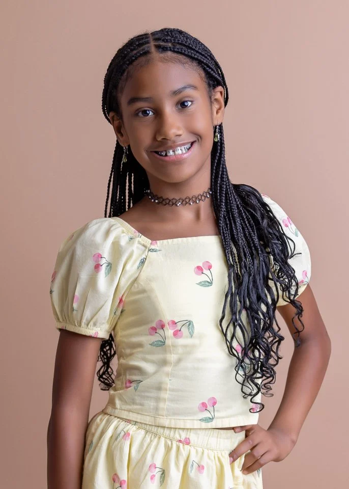 A young girl with long braided hair, smiling, wearing a yellow dress with pink cherry blossom print, a black choker, and earrings, standing against a beige background.