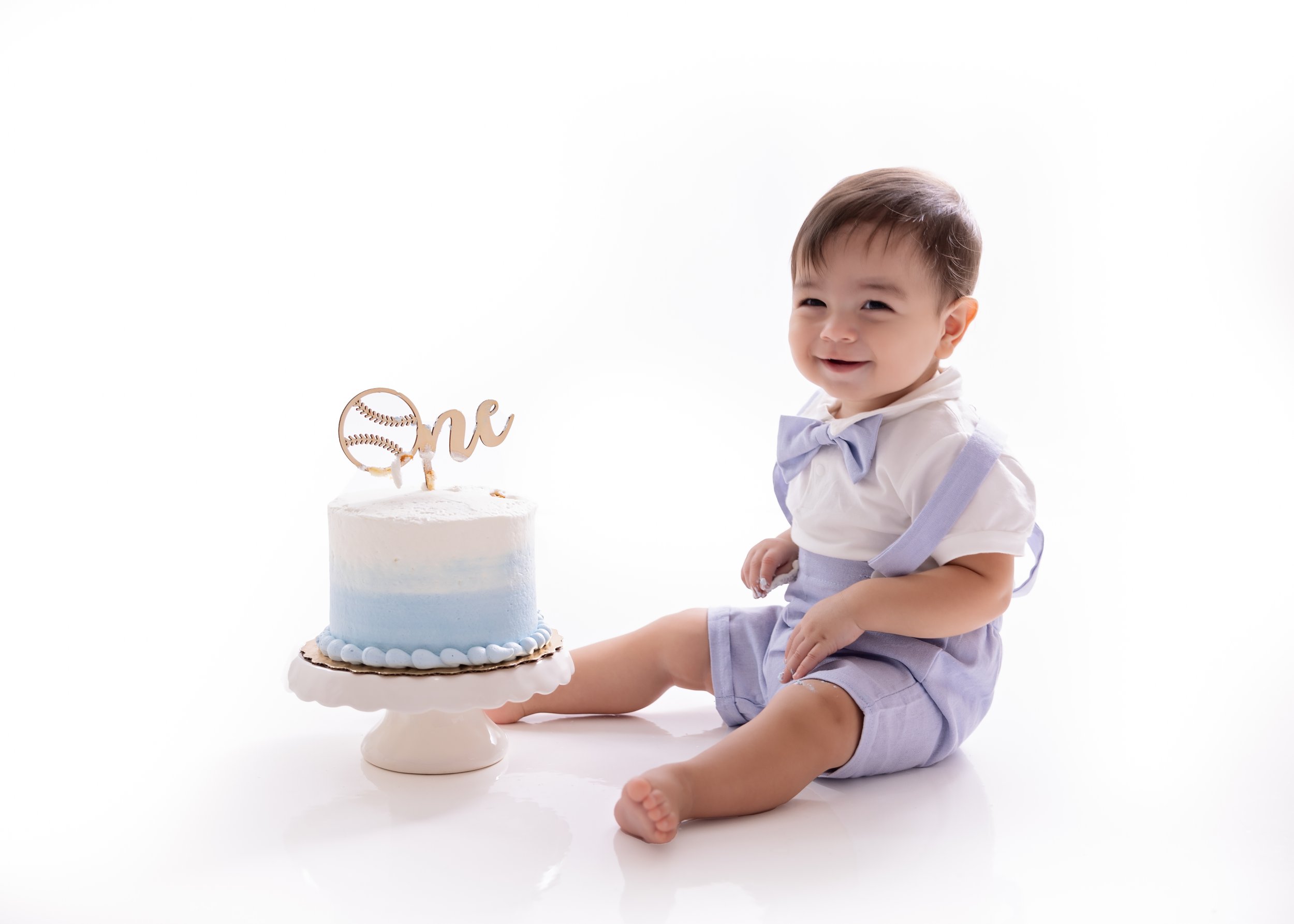 Smiling baby boy sitting next to a blue and white birthday cake with a "one" topper on a white background.