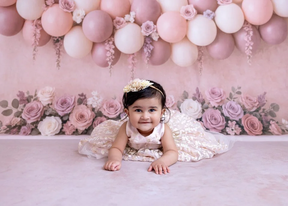 A smiling baby girl lying on her stomach on the floor, dressed in a cream-colored dress with a floral pattern and wearing a flower headband, in front of a pink floral and balloon backdrop.