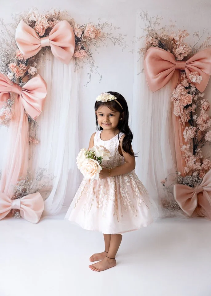 A young girl in a fancy white dress with lace details, holding a bouquet of light pink roses, standing in front of a decorated backdrop with large pink bows and pink flowers.