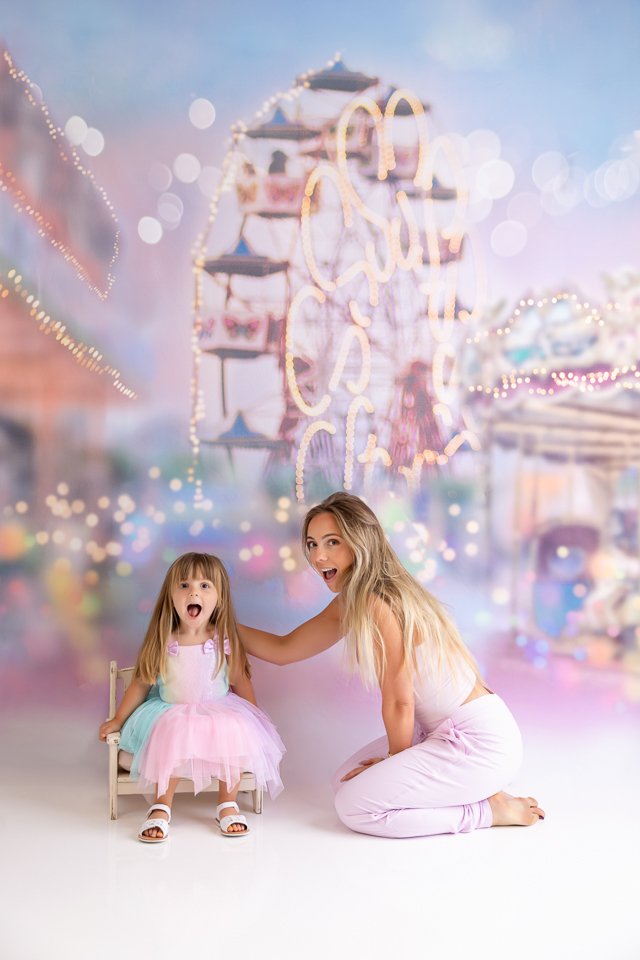 A woman and a young girl sitting on the floor in front of a colorful, blurred amusement park background, both with surprised and happy expressions.