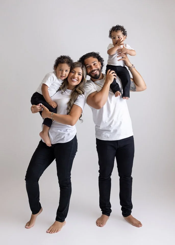 A happy family of four, including a mother, father, and two children, standing together against a plain white background. The mother is holding one child, and the father is holding the other child on his shoulders.