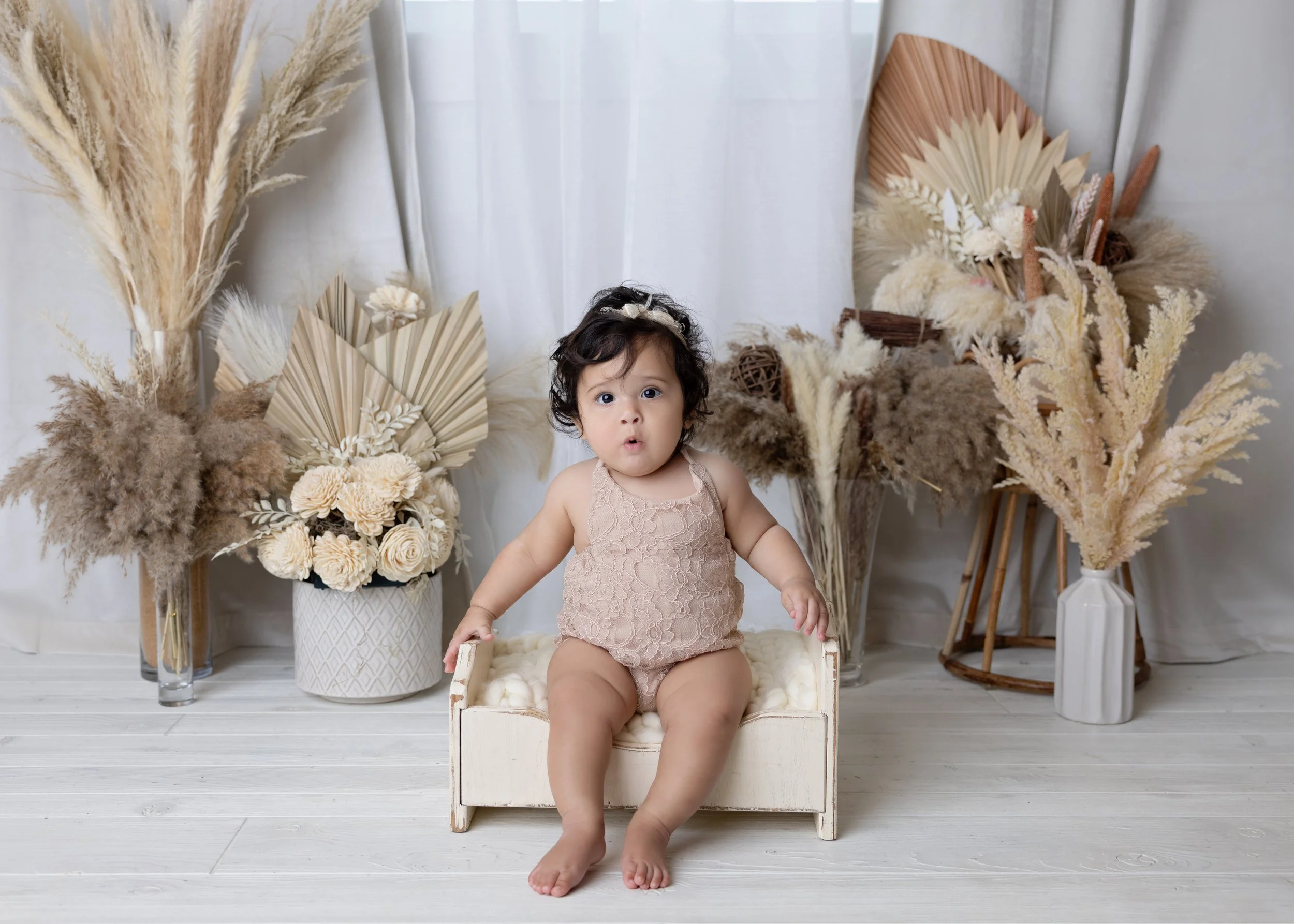 Young child sitting on small white wooden bed, surrounded by neutral-toned dried flower arrangements in vases, against light background with white curtains