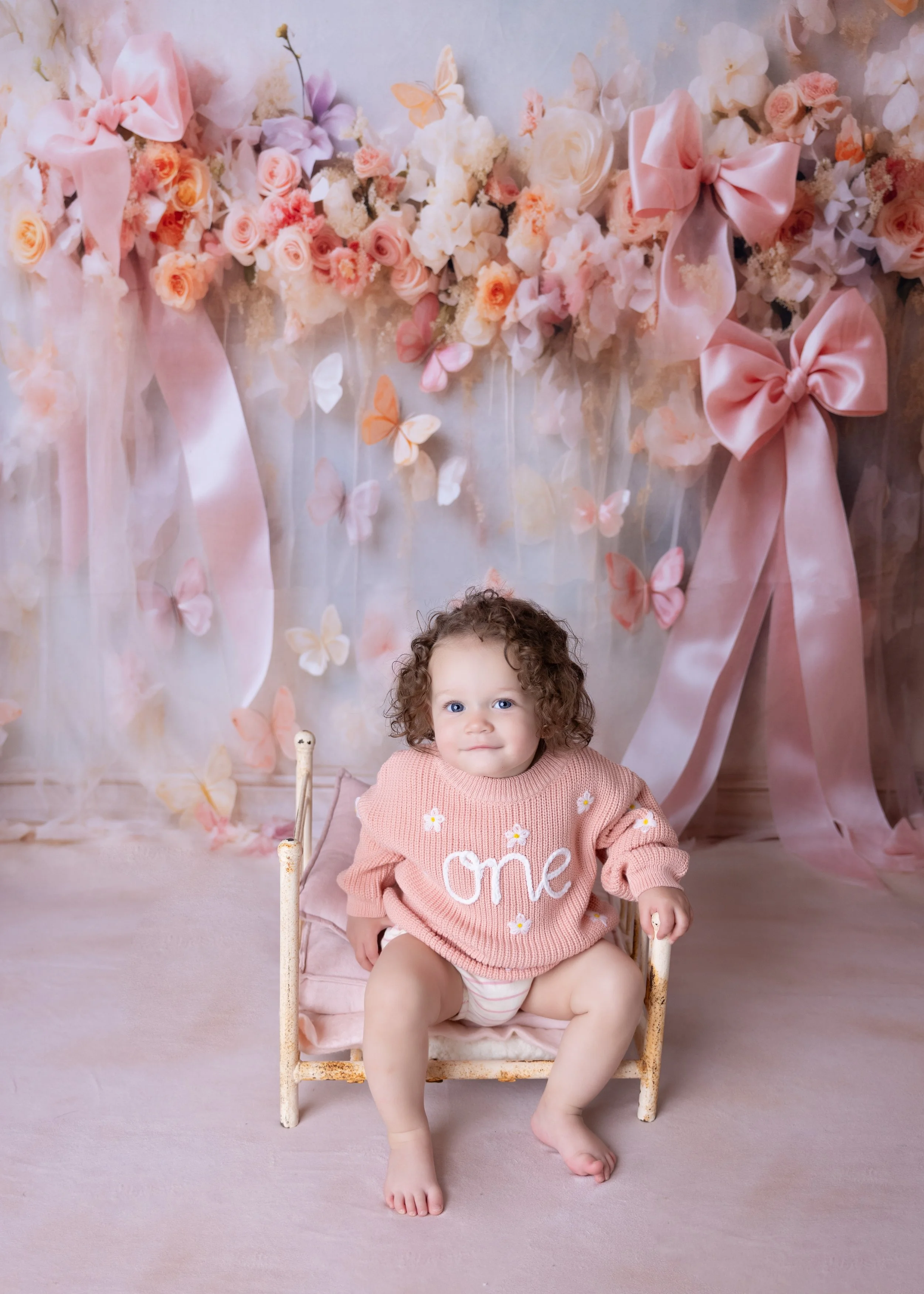 A young child with curly hair, wearing a pink sweater with 'one' written on it, sitting on a small pink chair in front of a floral backdrop decorated with pink and white flowers, butterflies, and large pink bows.